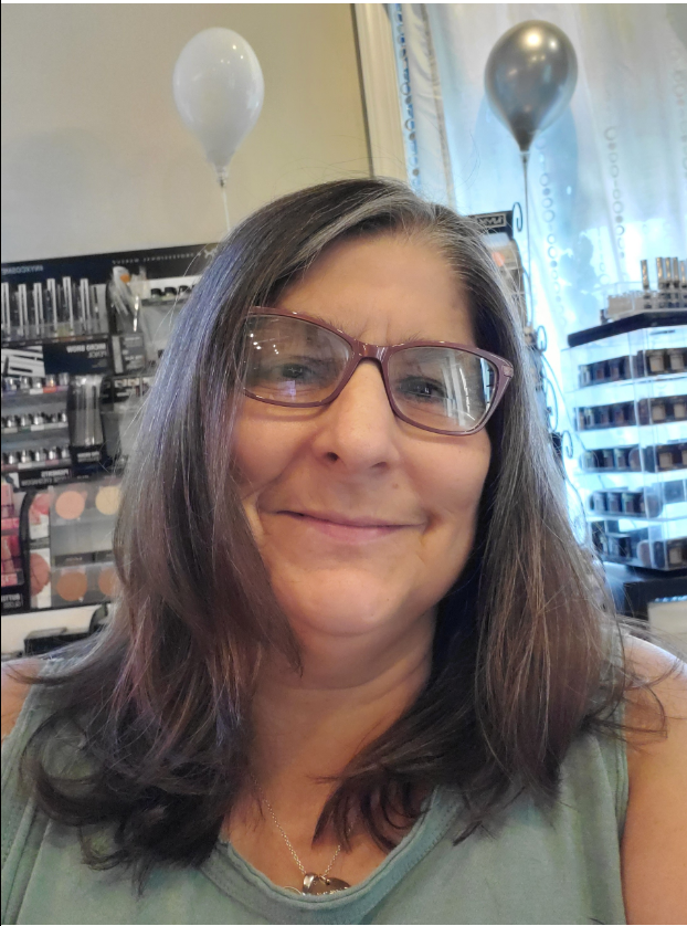 A woman with long dark hair and glasses smiling in a makeup store, with balloons and various makeup products behind her.