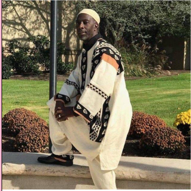 Man wearing traditional African attire with a patterned jacket, white robe, and white head covering, standing outdoors on a sidewalk with flowers and trees in the background.