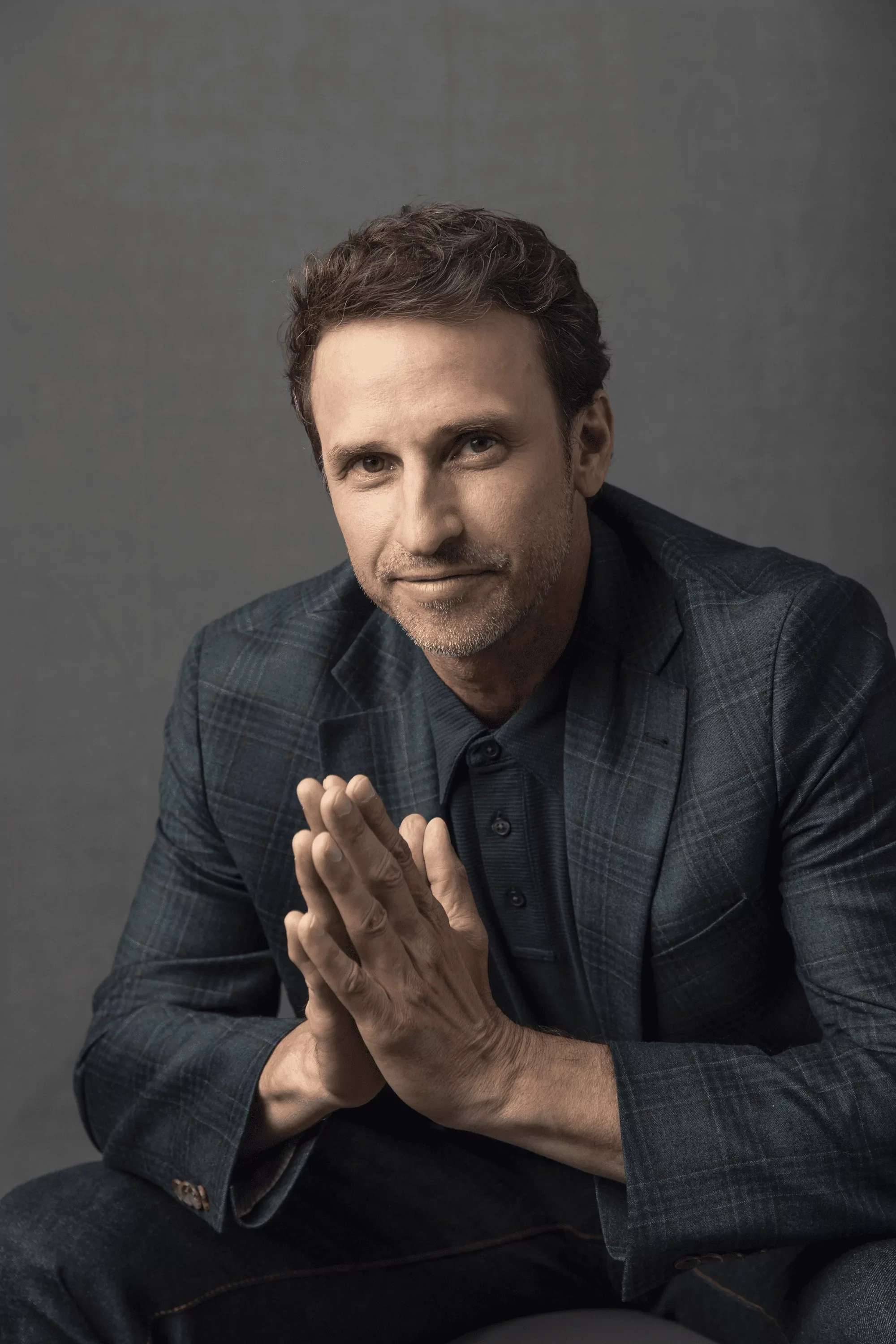 Jeff Salzenstein in a checkered blazer and dark shirt sitting with hands pressed together, looking at the camera with a neutral expression against a gray background.