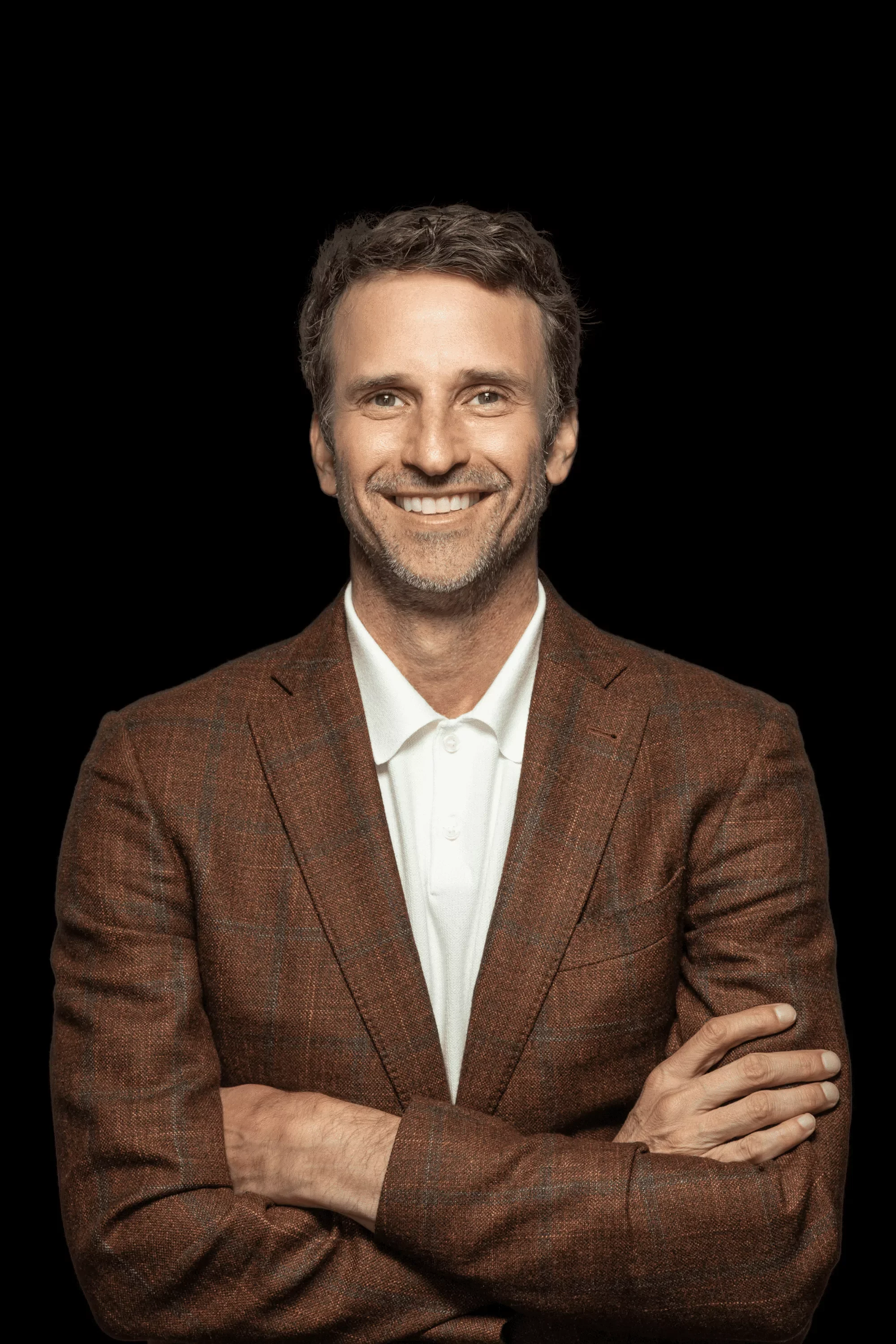 A smiling Jeff Salzenstein with short gray hair and beard, wearing a brown checked blazer and white collared shirt, standing against a black background.