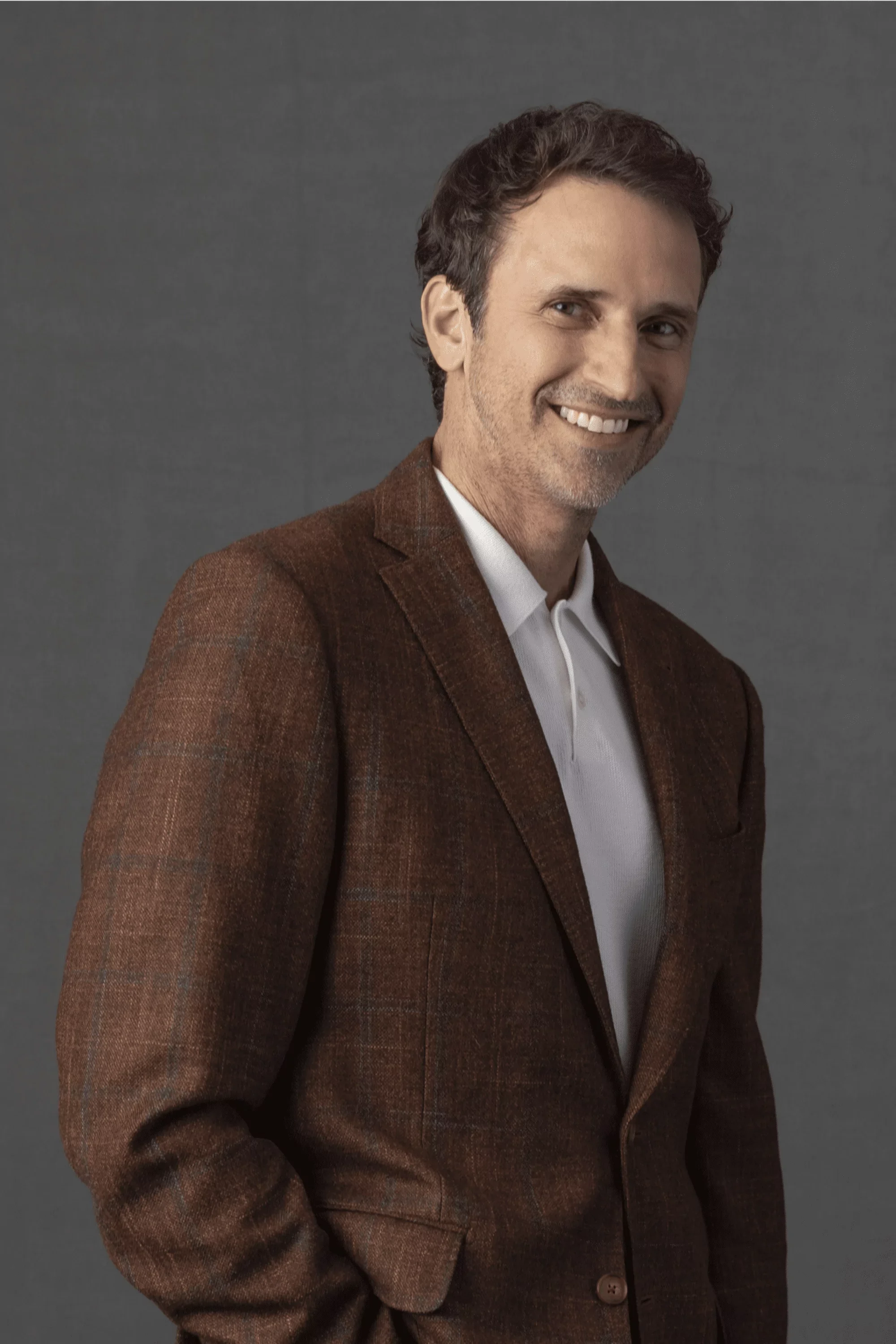 Portrait of Jeff Salzenstein in a brown checked blazer and white shirt, smiling, standing against a plain gray background.