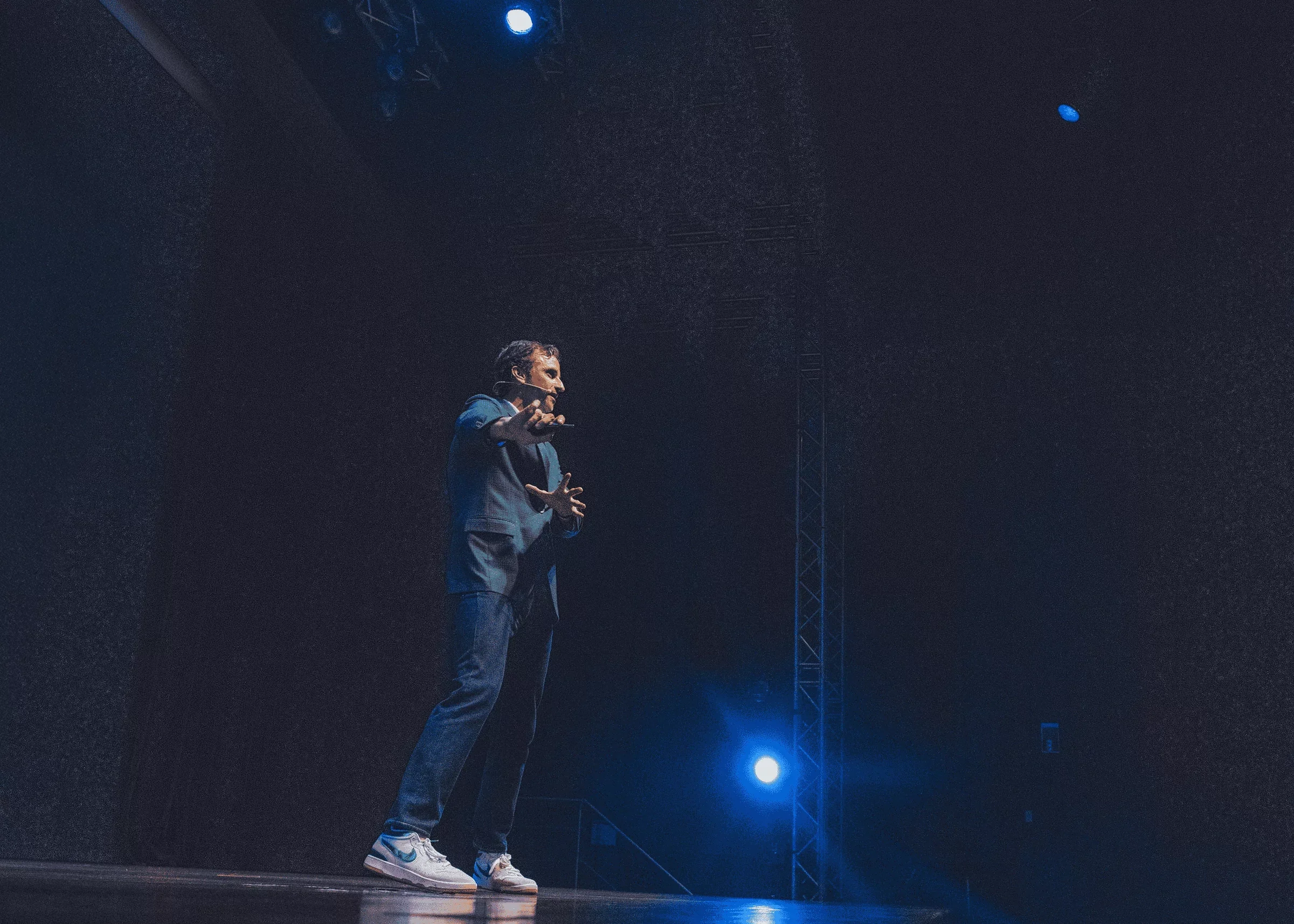 Jeff Salzenstein on stage delivering a presentation or speech, illuminated by blue stage lighting, with a dark background and visible metal truss structure.