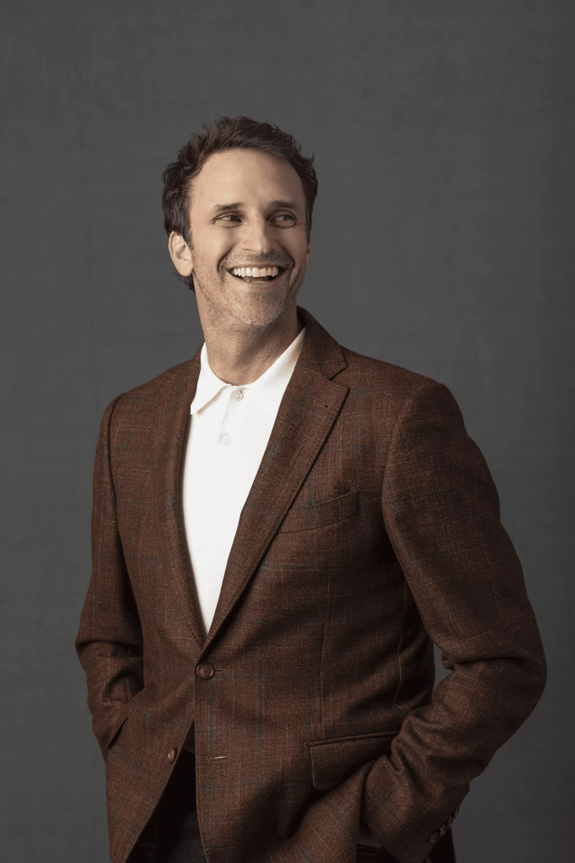 Jeff Salzenstein with dark curly hair and light skin, wearing a brown textured blazer over a white shirt, posing against a plain dark gray background.