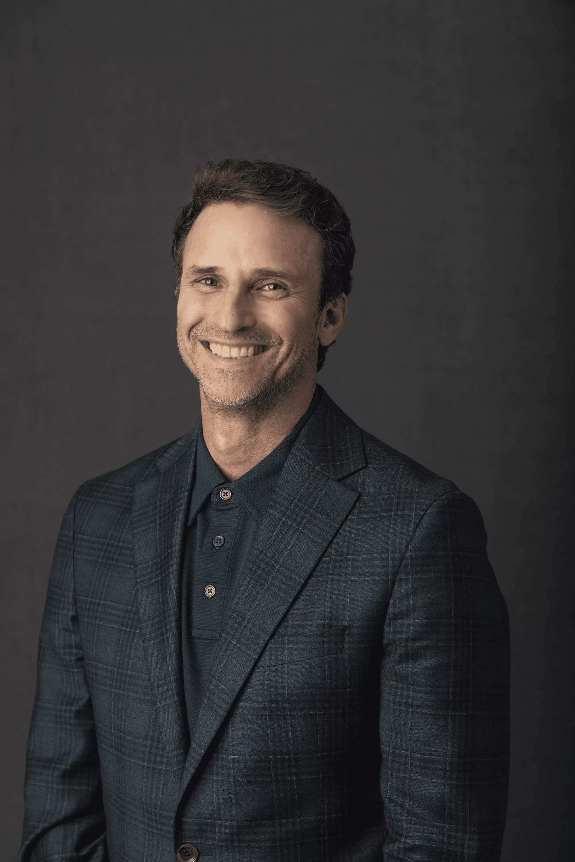 A smiling Jeff Salzenstein with gray hair and a beard, wearing a dark plaid blazer over a navy shirt, standing against a dark background.