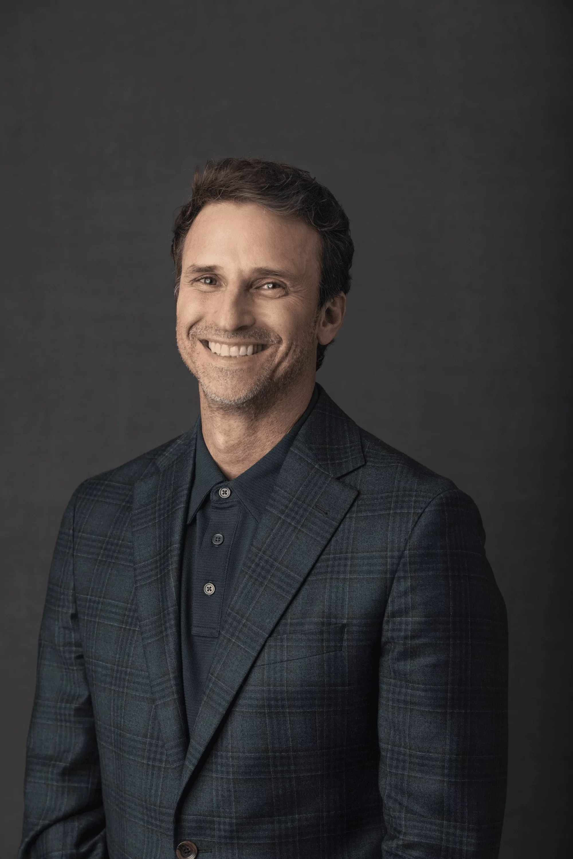 Portrait of Jeff Salzenstein in a dark plaid blazer and navy shirt against a gray background.