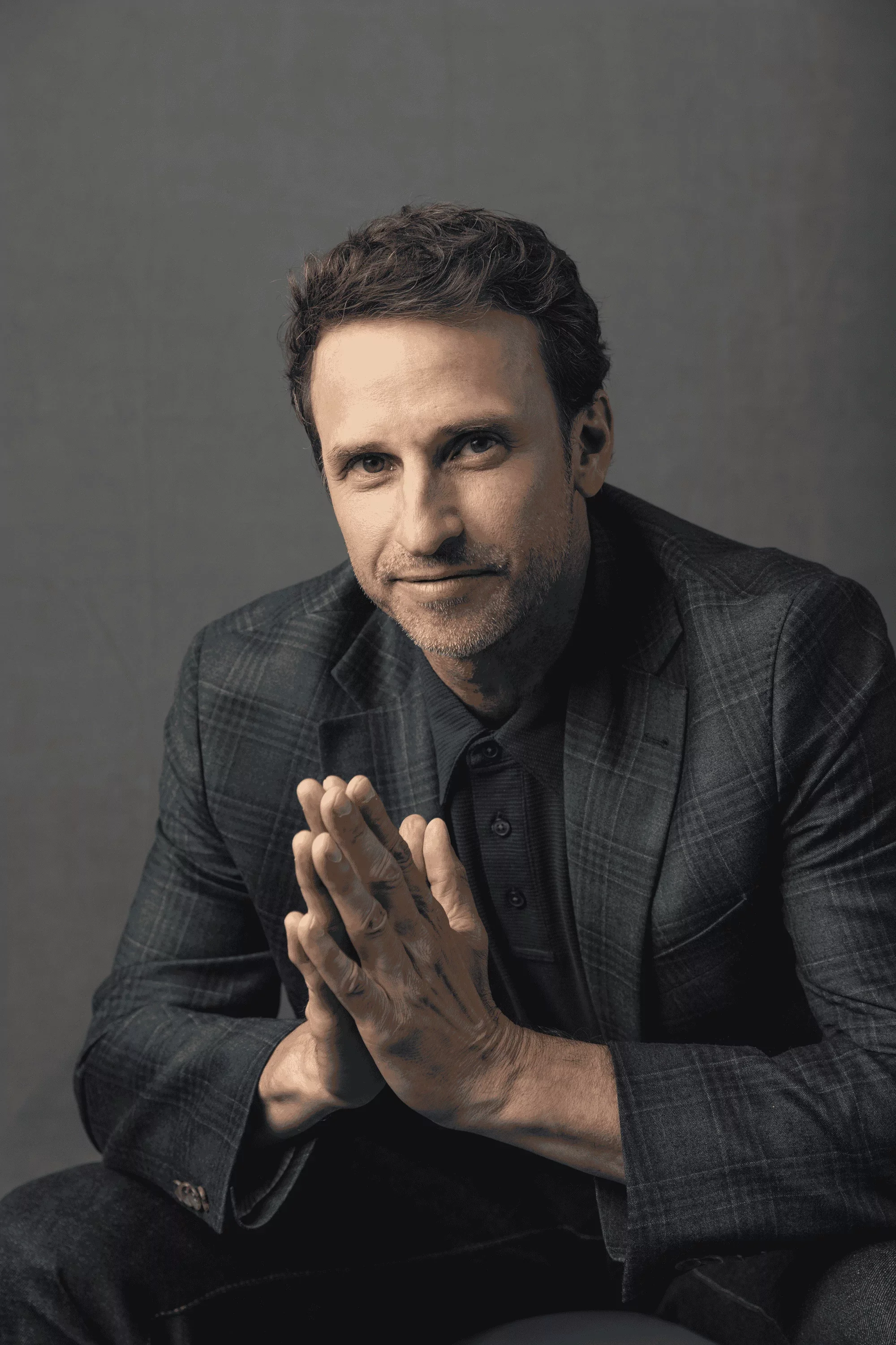 Jeff Salzenstein in a dark plaid blazer and black shirt, sitting with his hands clasped together, smiling slightly at the camera.