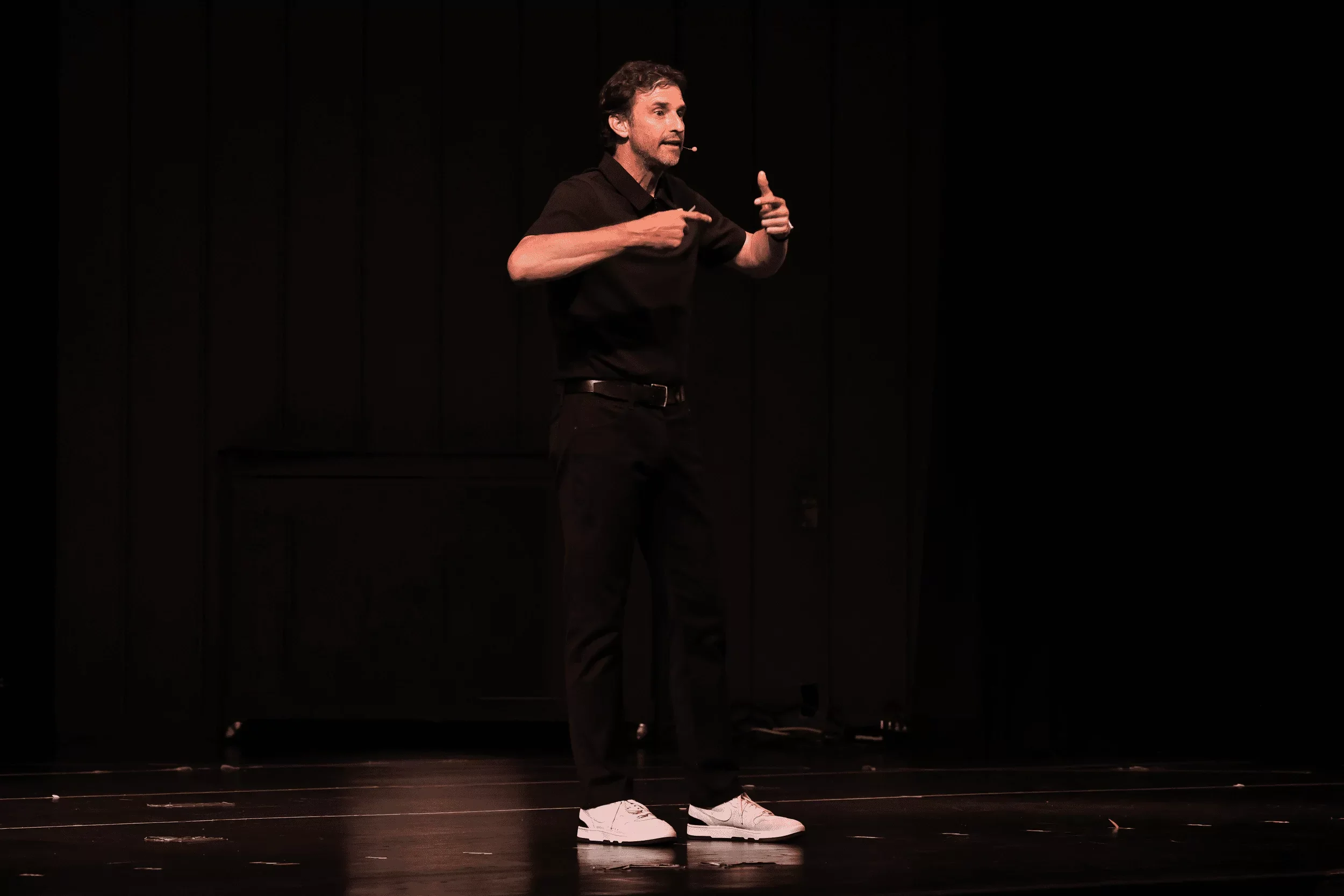 Jeff Salzenstein in a black shirt and pants standing on a stage with a black background, gesturing with his hands.