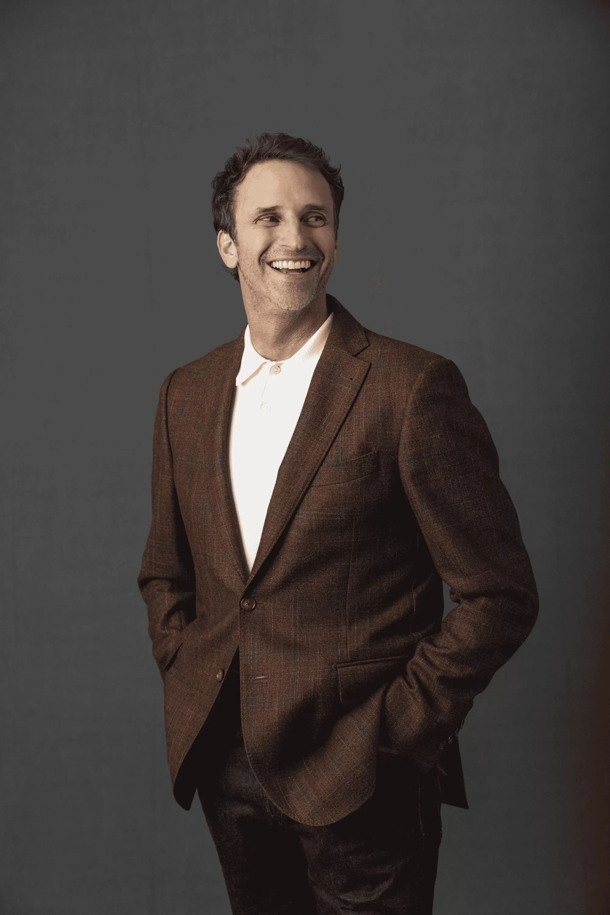 Jeff Salzenstein with dark, slightly curly hair and light skin wearing a brown plaid suit and white shirt, smiling and looking to his left, against a plain dark gray background.