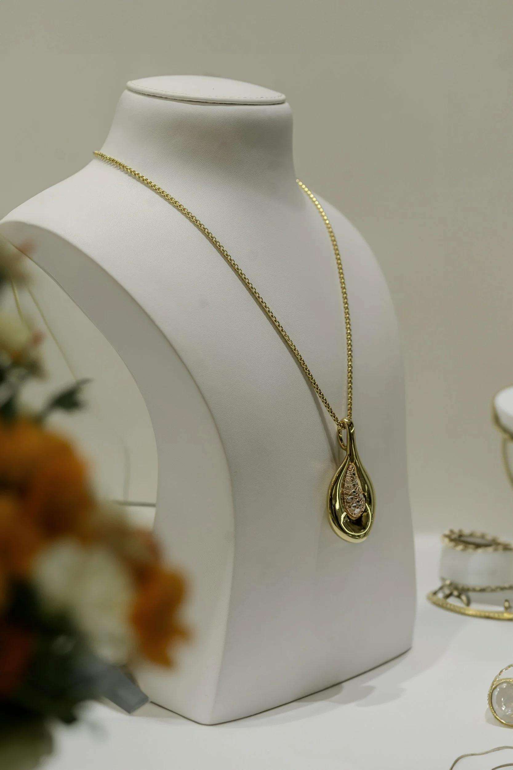 Gold necklace with teardrop-shaped pendant displayed on a white jewelry bust, with blurred flowers in the foreground.