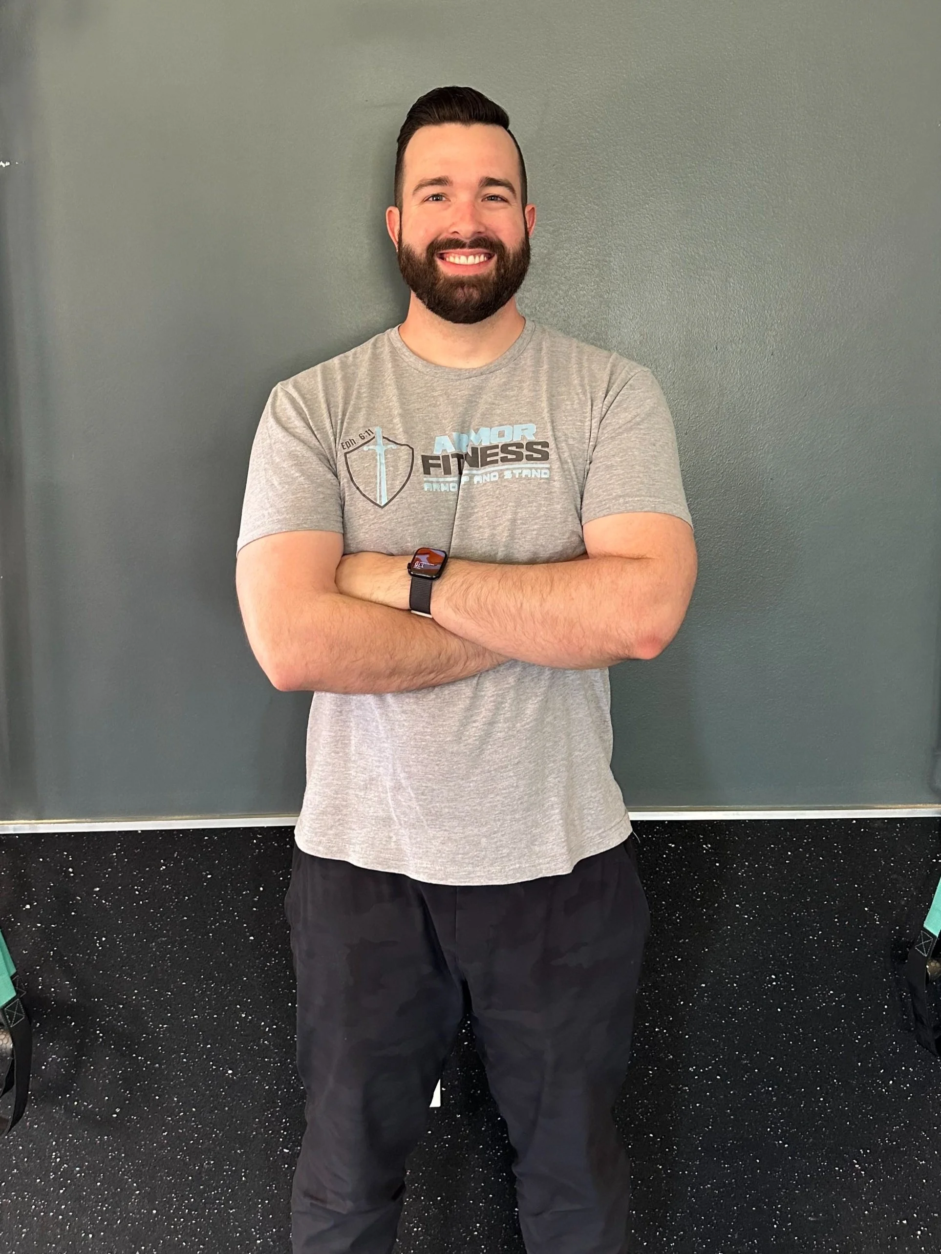 A man with a beard and short hair wearing a gray T-shirt and black pants stands smiling with arms crossed against a gray wall.