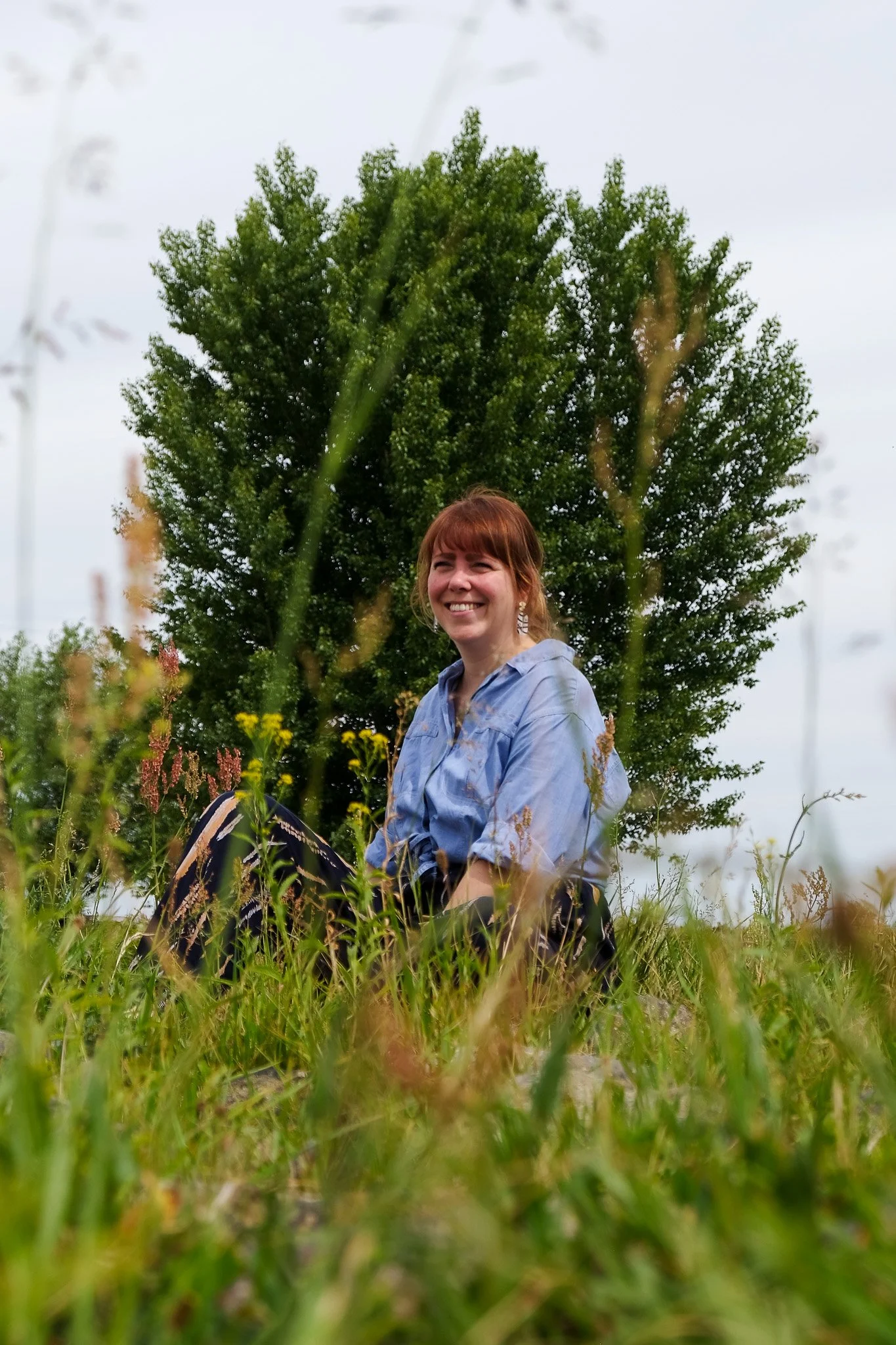 Vrouw zit in een grasveld met een boom op de achtergrond, lachend en genietend van de natuur.
