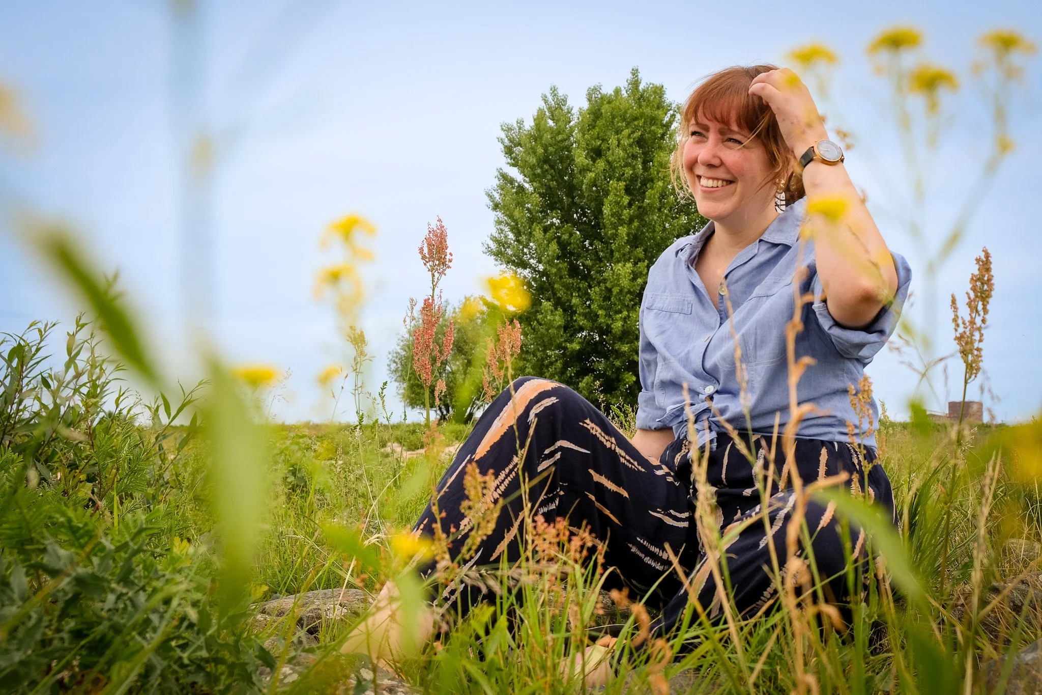 Vrouw zit in een veld met groen gras en bloemen, lachend onder een blauwe lucht, met een boom op de achtergrond.