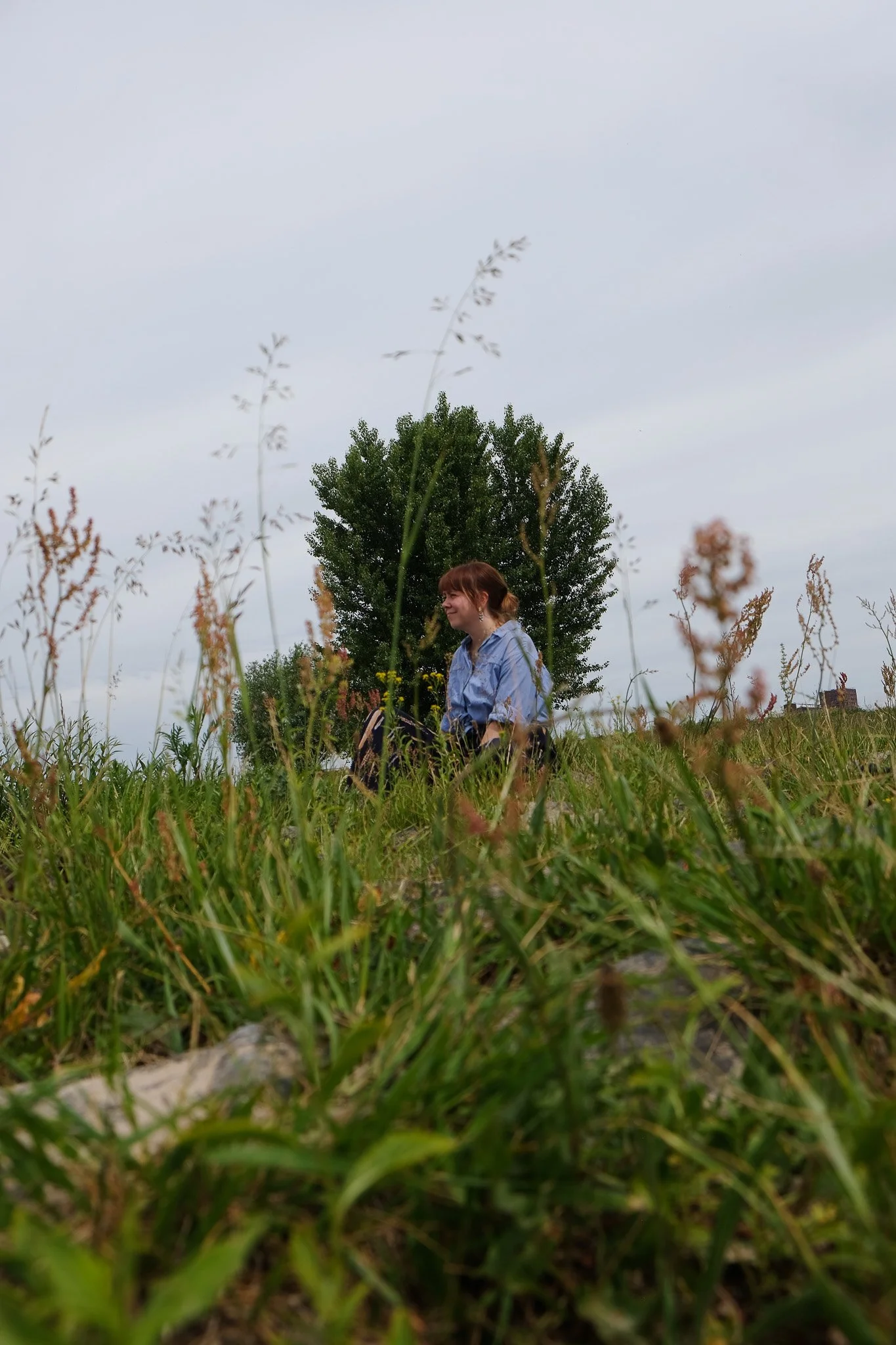 Een vrouw met rood haar in een blauwe blouse knielt in een grasveld met een grote boom op de achtergrond onder een bewolkte hemel.