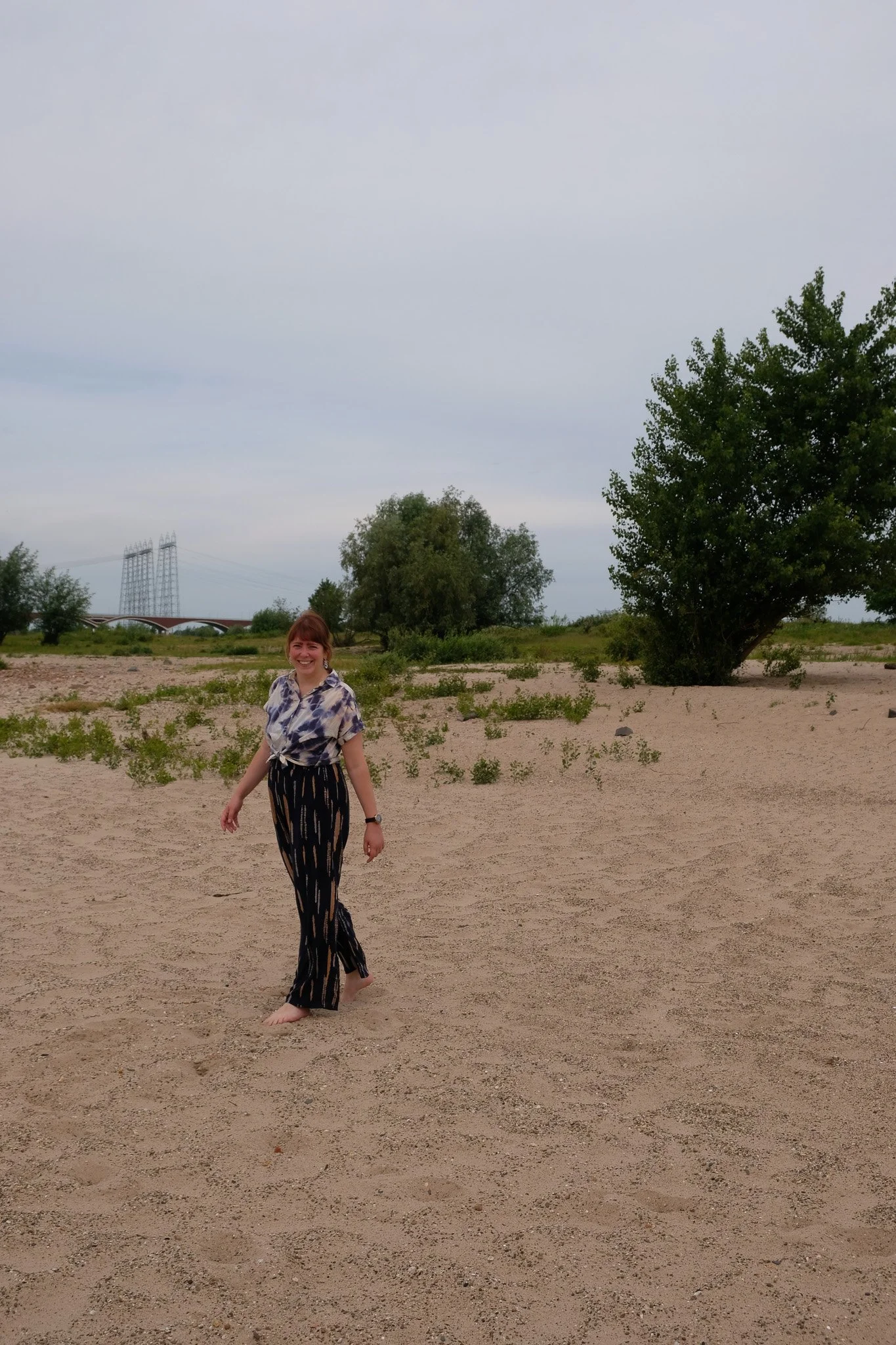 Vrouw die op een zandstrand wandelt, glimlachend naar de camera, met bomen en een brug op de achtergrond.