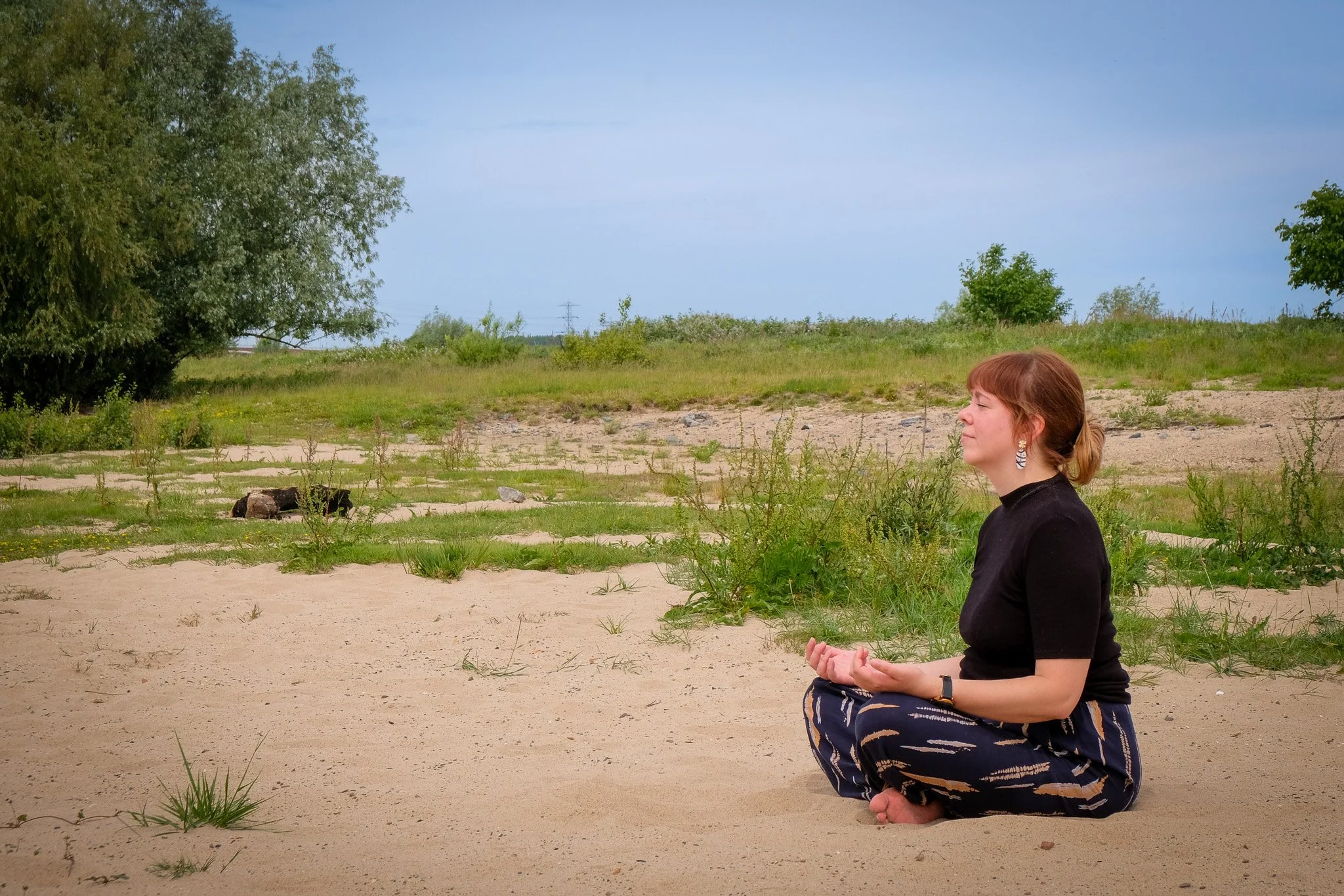 Vrouw die in lotushouding op het zand zit en mediteert op een natuurlijke plek met gras en bomen.