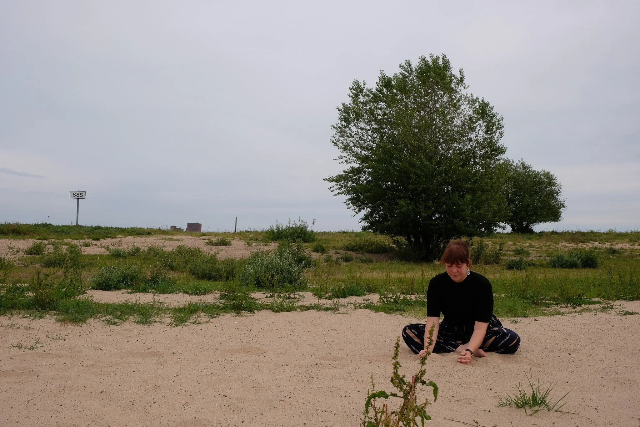 Vrouw zit in plooi op zand bij een groene boom onder een bewolkte hemel.