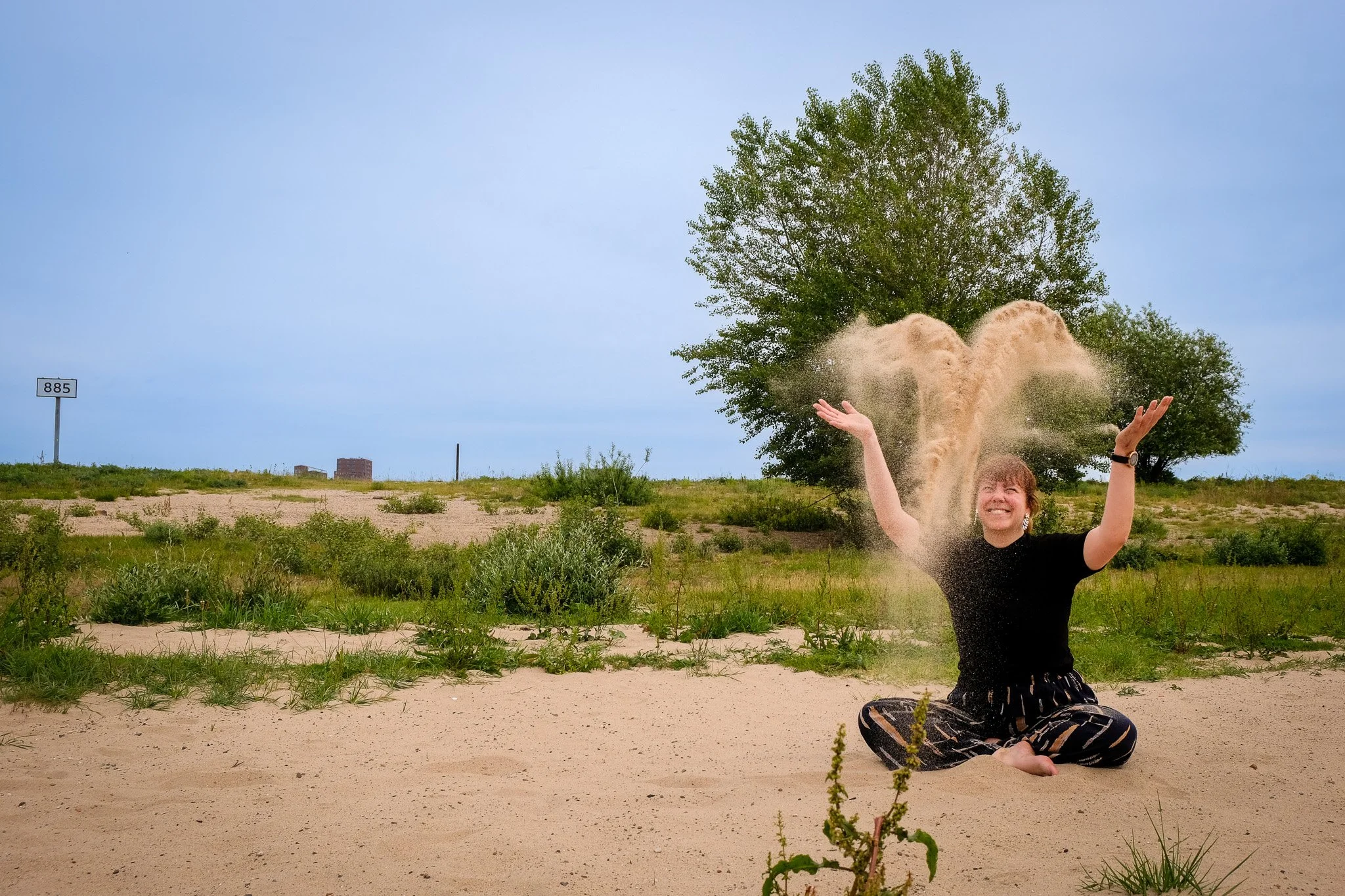 Vrolijke vrouw zittend op zand met zand dat omhoog waait, onder een grote boom en een heldere blauwe lucht.