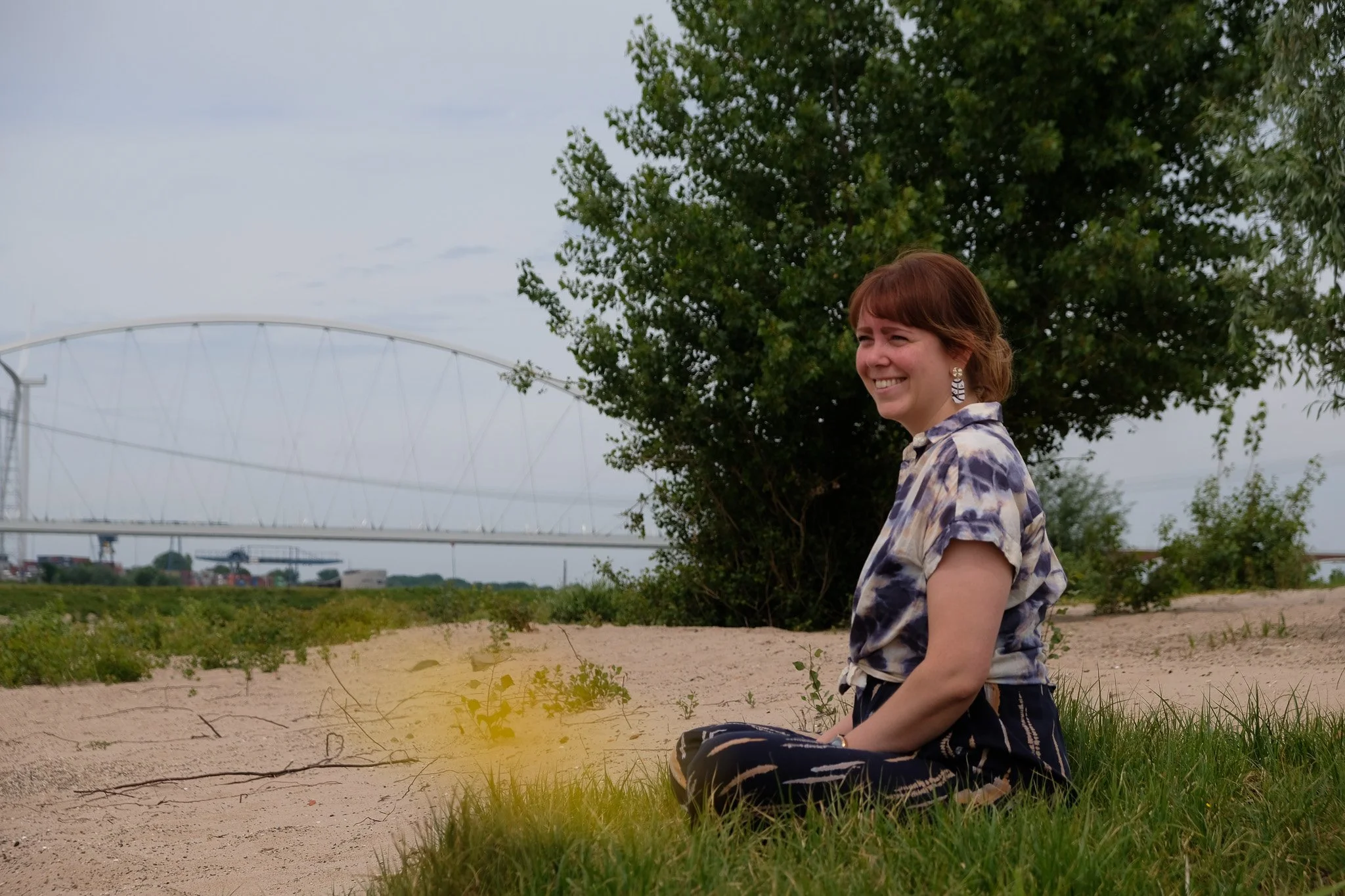 Een lachende vrouw zit buiten op het gras onder een boom met een brug op de achtergrond.