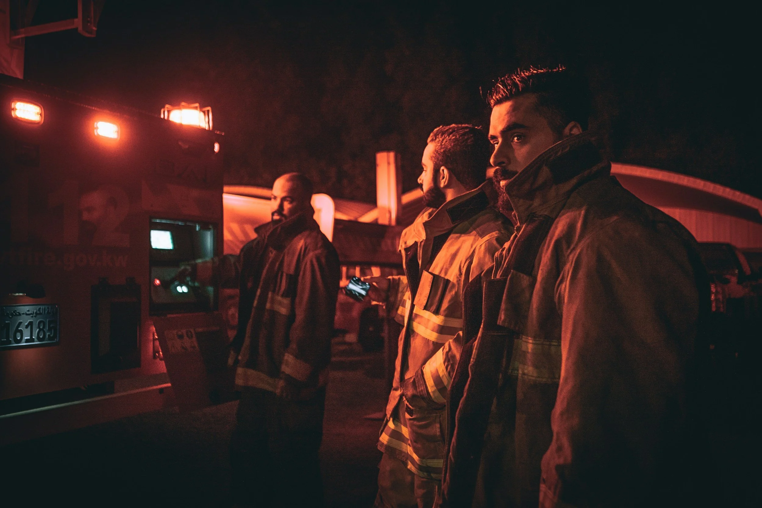 Firefighters in protective gear near a fire truck at night, lit by red emergency lights.
