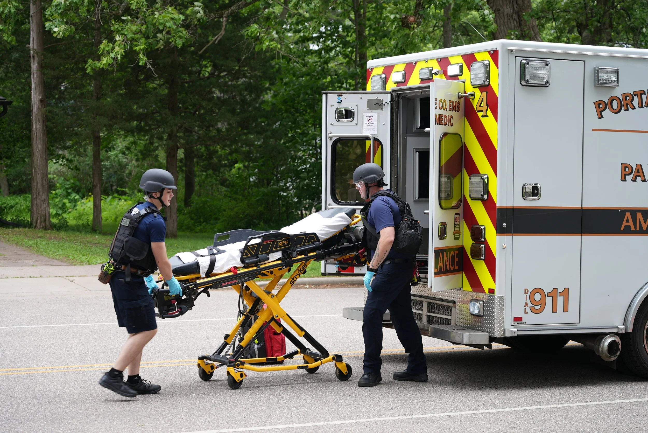 Two emergency responders wearing helmets and gloves are loading a patient on a stretcher into an ambulance on a street with green trees in the background.