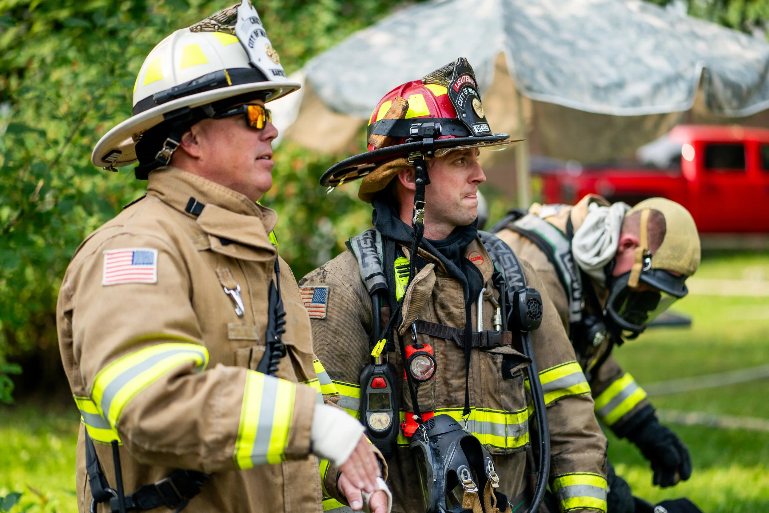 Three firefighters wearing protective gear and helmets, standing outdoors with a grass background, focused and prepared for action.