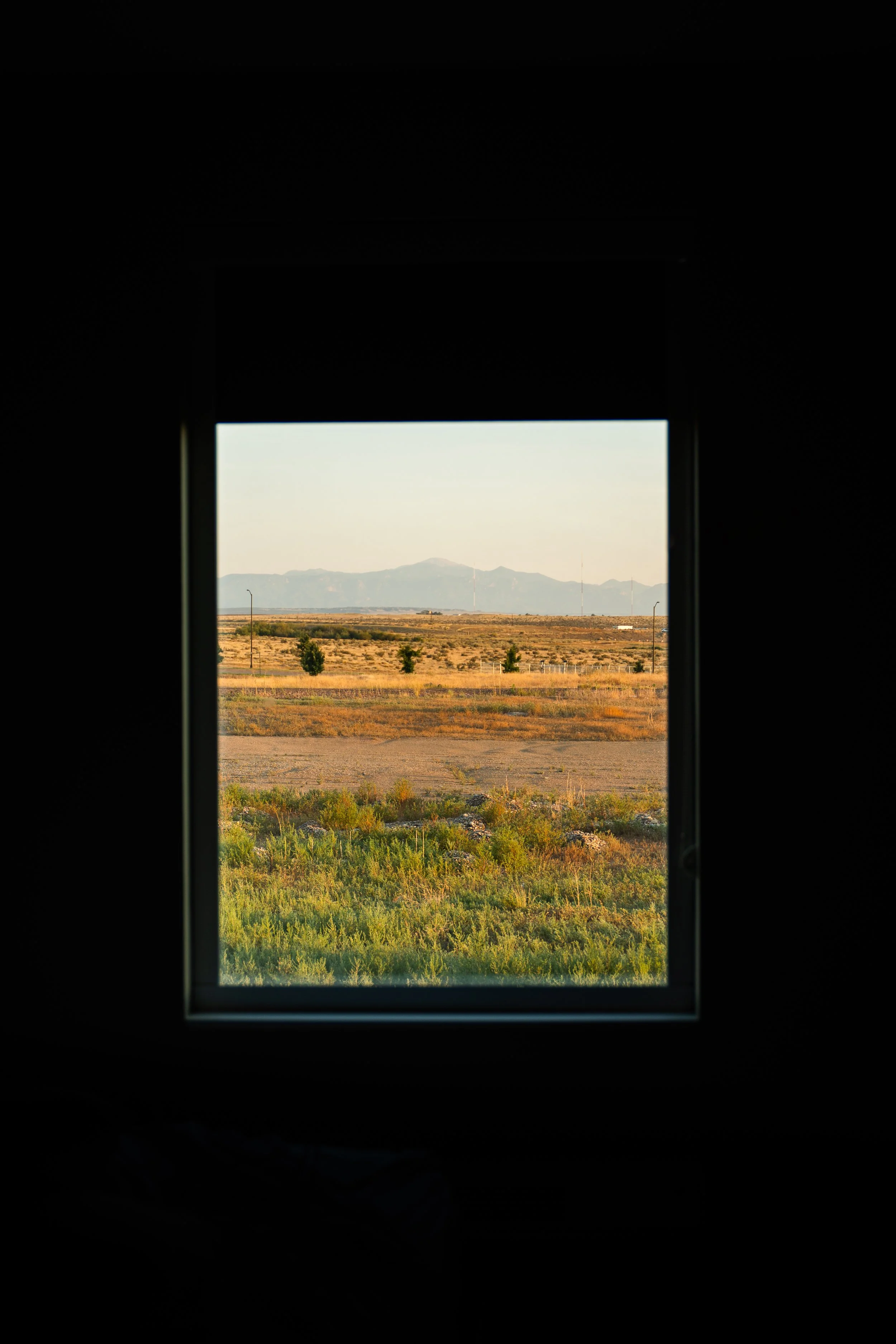 View of a landscape with mountains in the distance, fields, and scattered trees, seen through a window.
