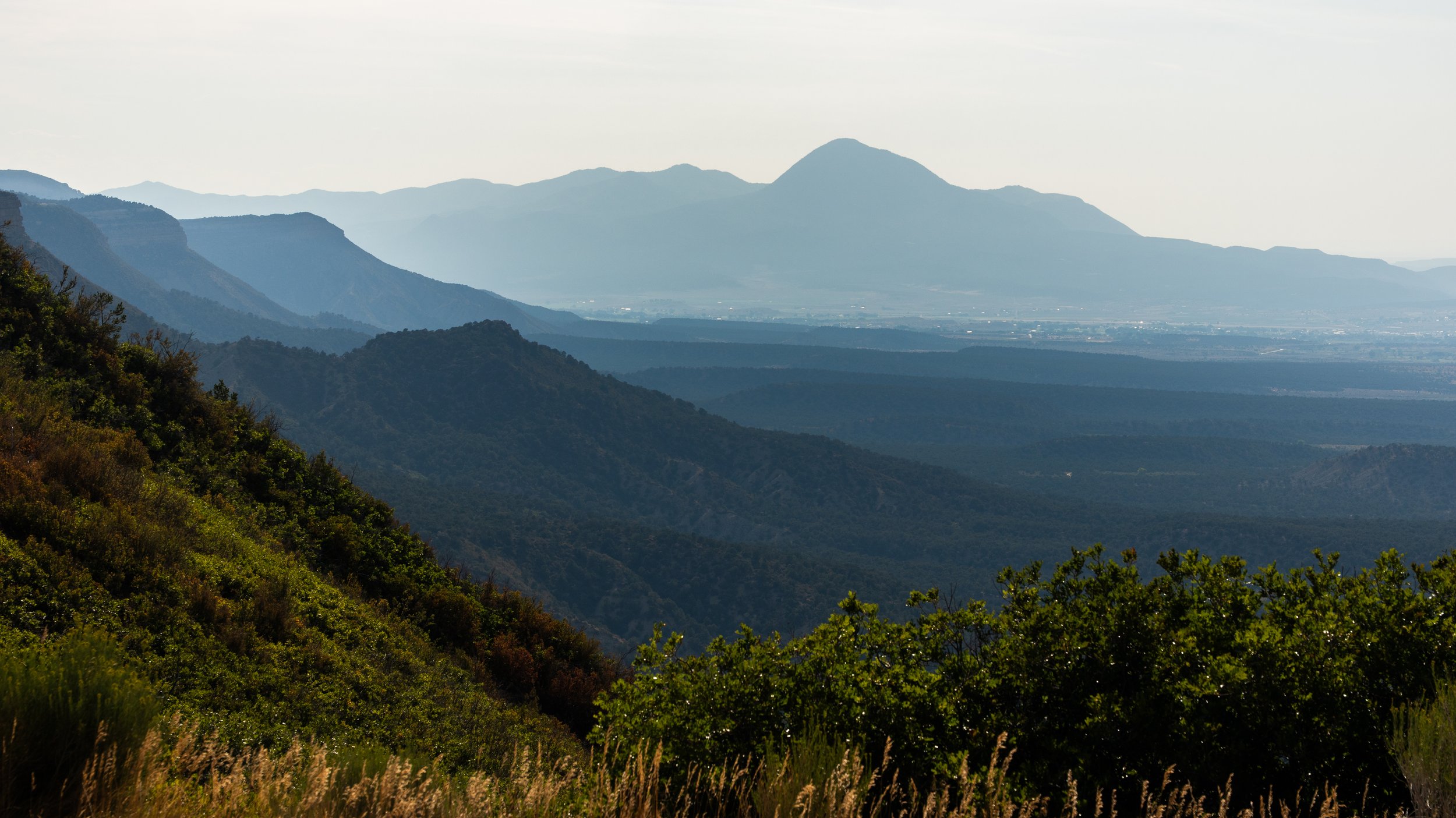 Landscape view of rolling green hills with layers of mountains in the distance, under a pale cloudy sky.