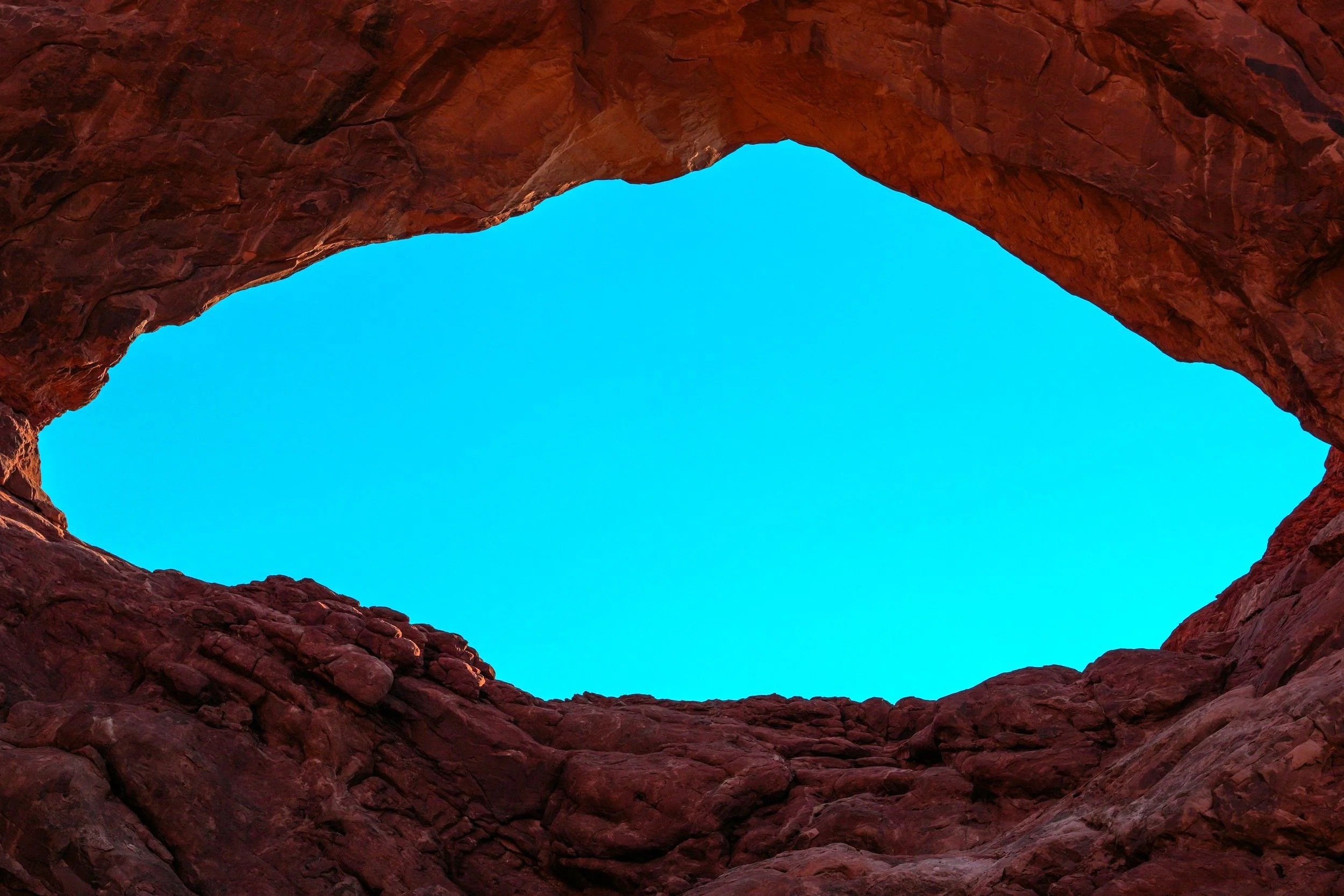 Looking up from inside a red rock formation toward the bright blue sky through an open space in the rock.
