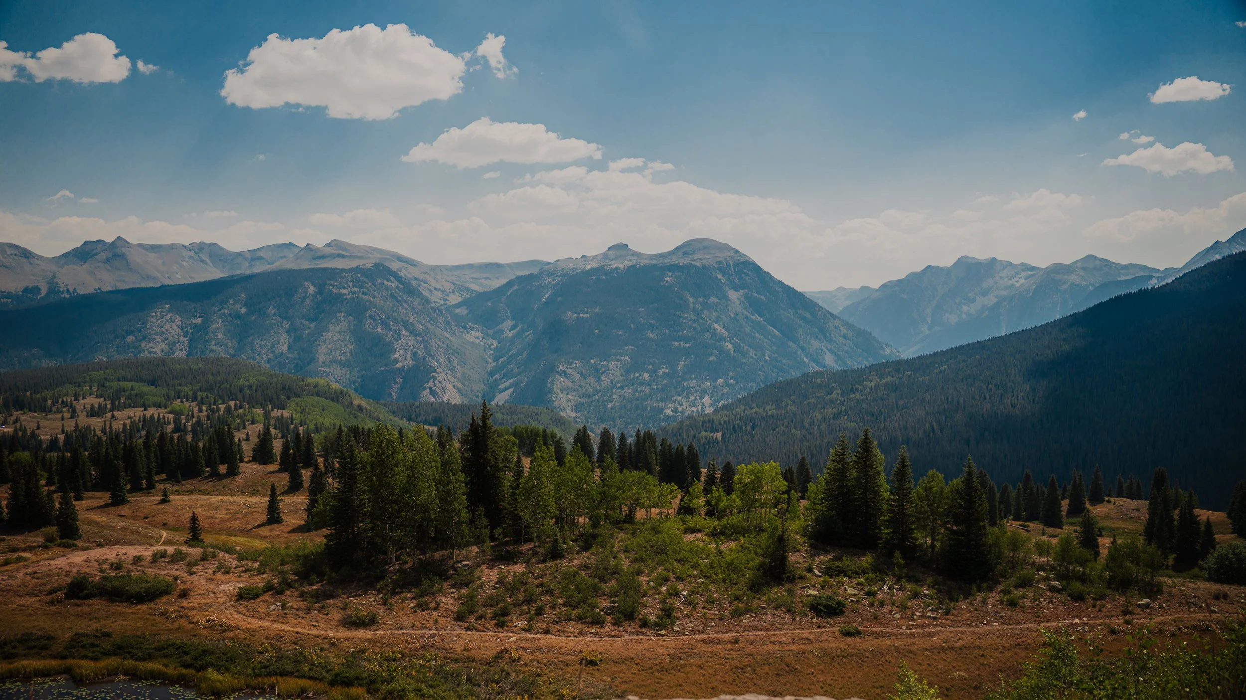 A scenic view of a mountain range under a blue sky with scattered clouds, forested hills, and a dirt path in the foreground.
