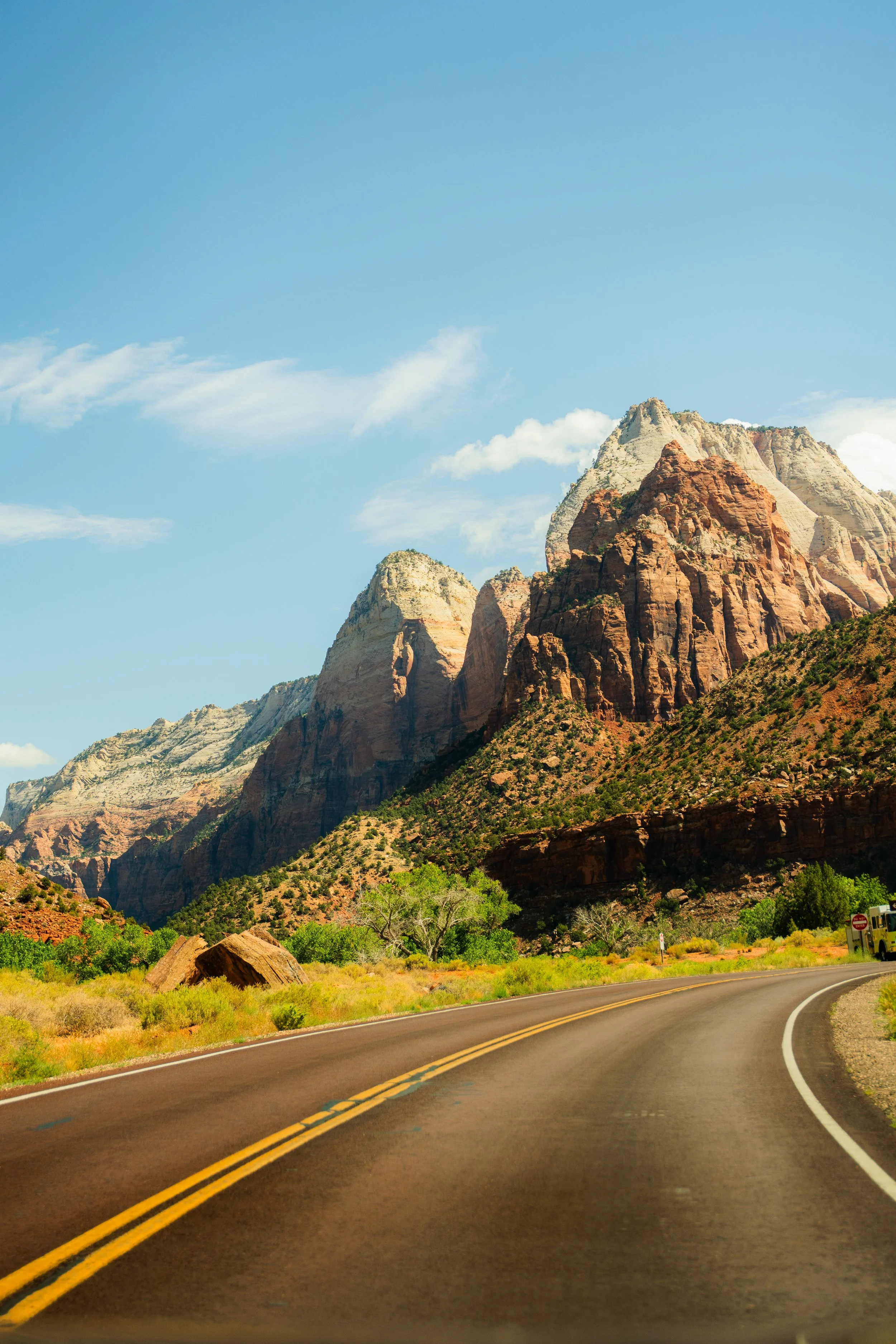 A winding road through a canyon with tall, rugged mountains under a bright blue sky with a few clouds.