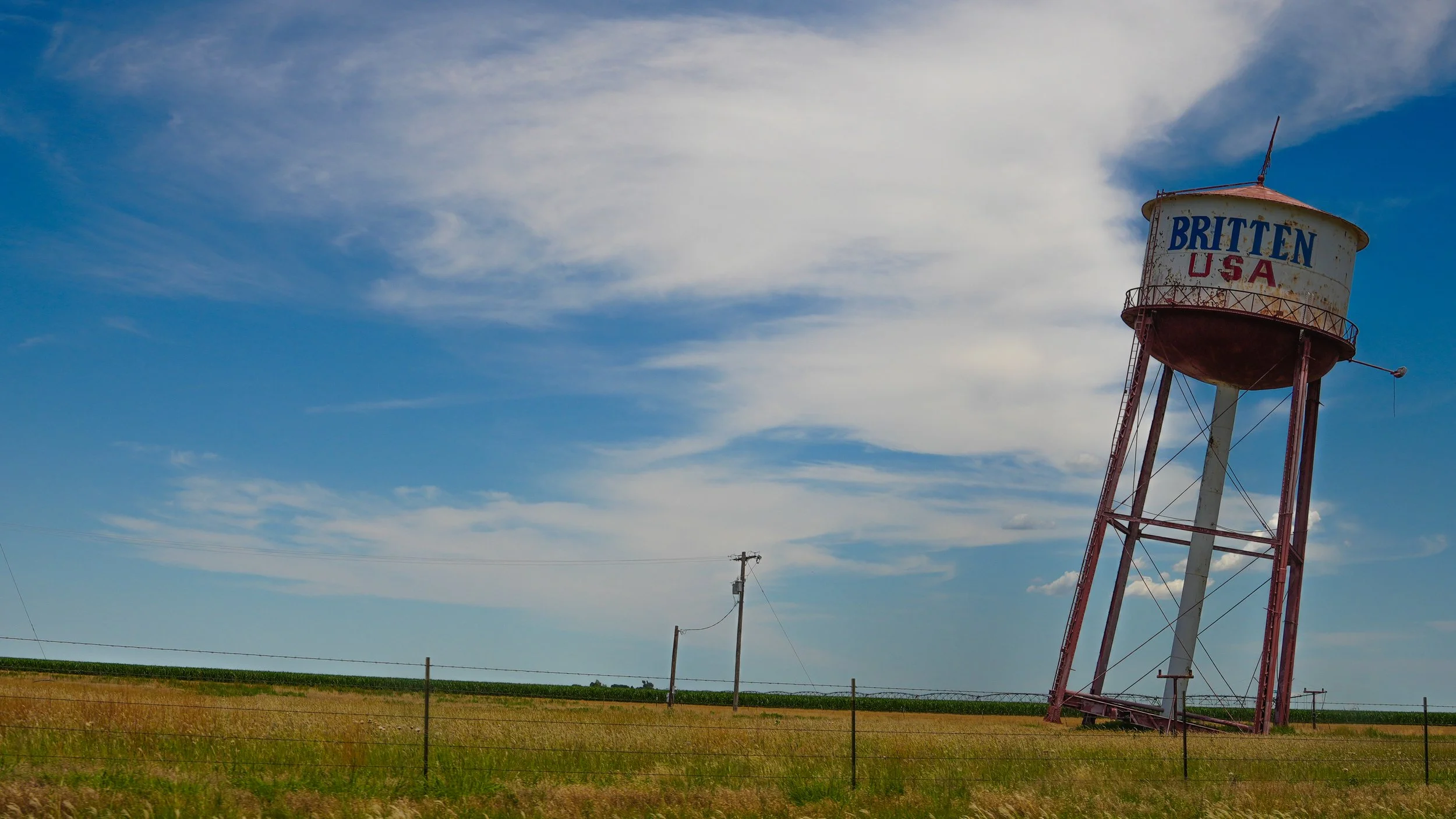 A rusty water tower with the words 'Britten USA' on it in a rural field under a partly cloudy sky.
