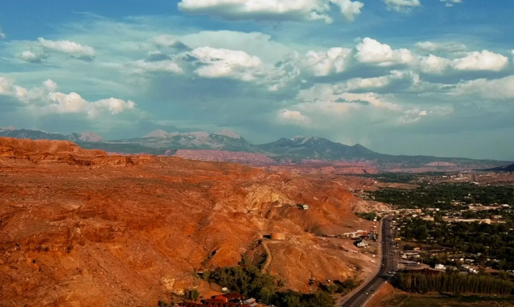 Scenic view of red and orange rocky hills with a winding road and a small town, mountains with snow-capped peaks in the background, and clouds in the sky.
