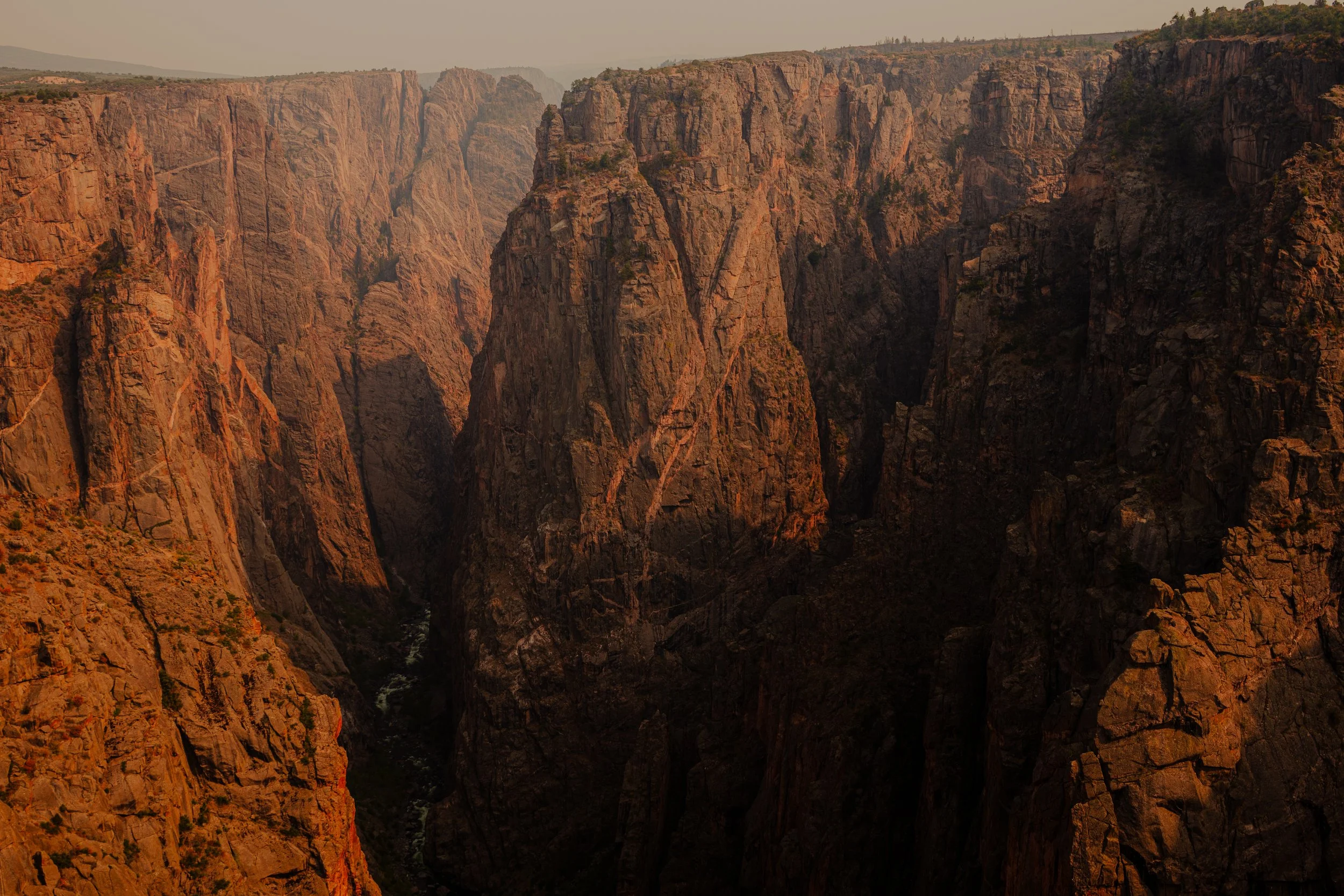 A view of a deep canyon with tall, rugged rock formations and cliffs, with a narrow river at the bottom, during sunset or sunrise.