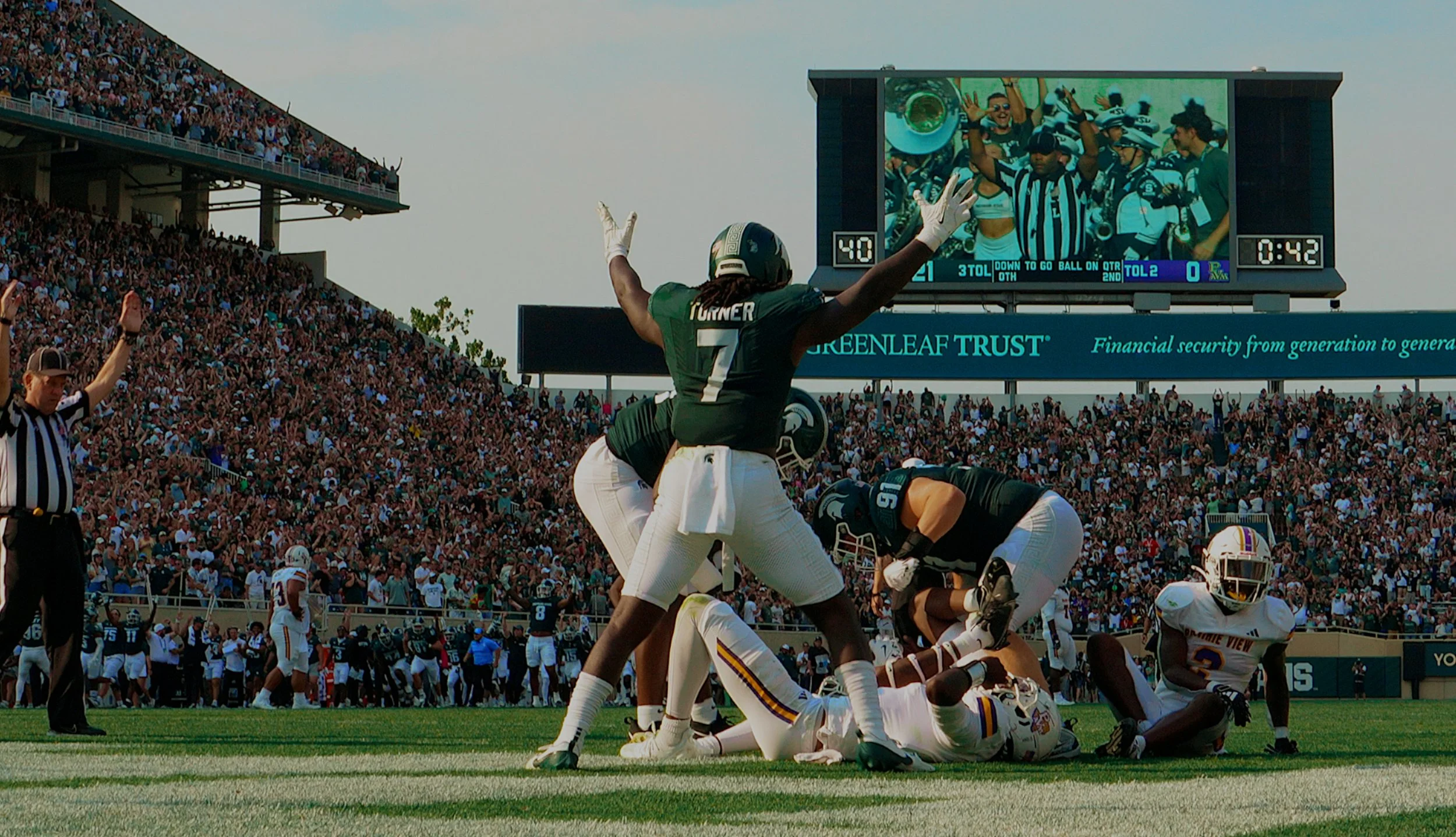 An American football game with players celebrating a touchdown or a significant play, with a large crowd in the stands and a large scoreboard displaying game details.
