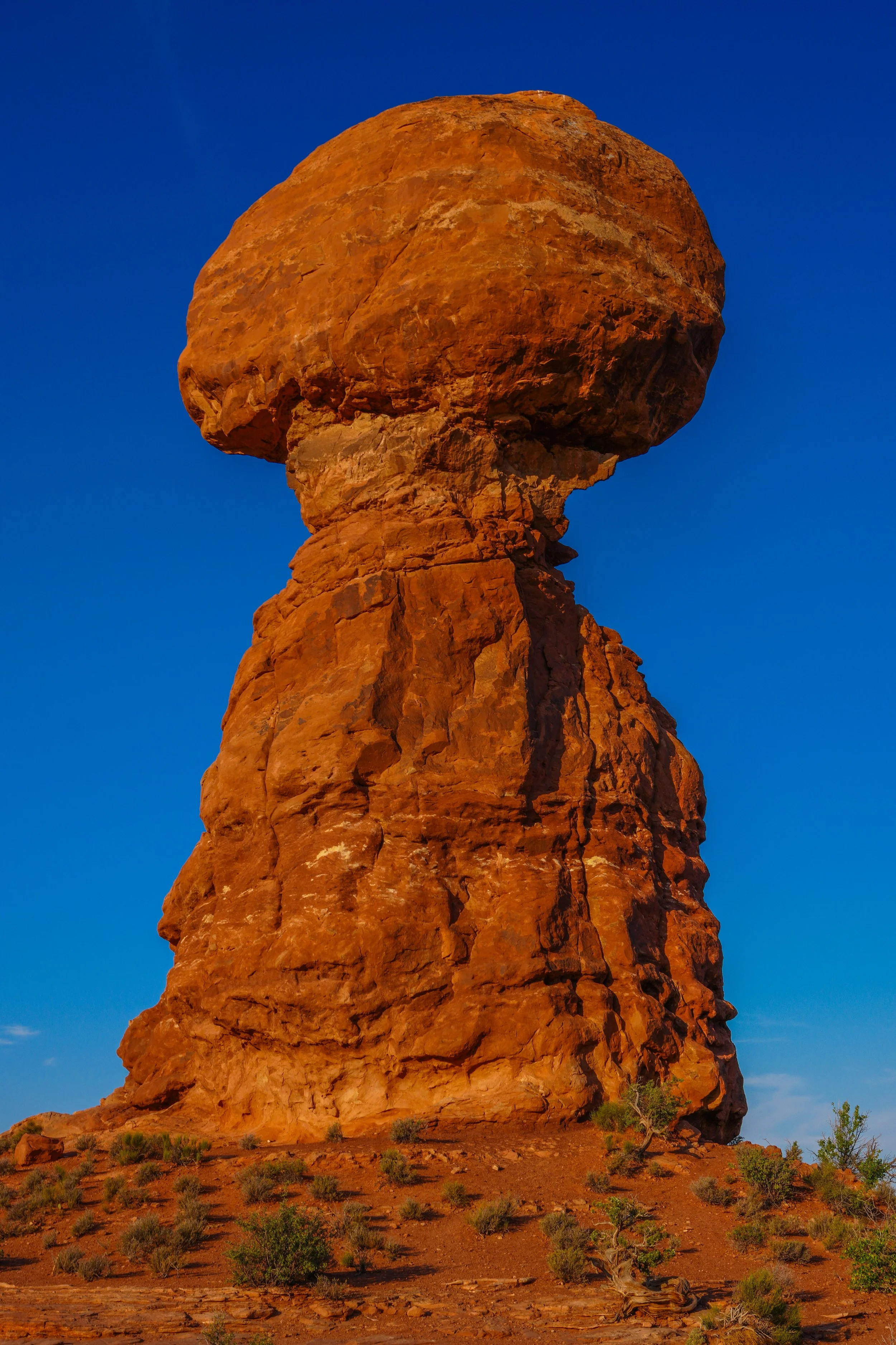 A large, reddish-brown rock formation with a giant boulder balanced on top, set against a clear blue sky in a desert landscape with sparse green vegetation.