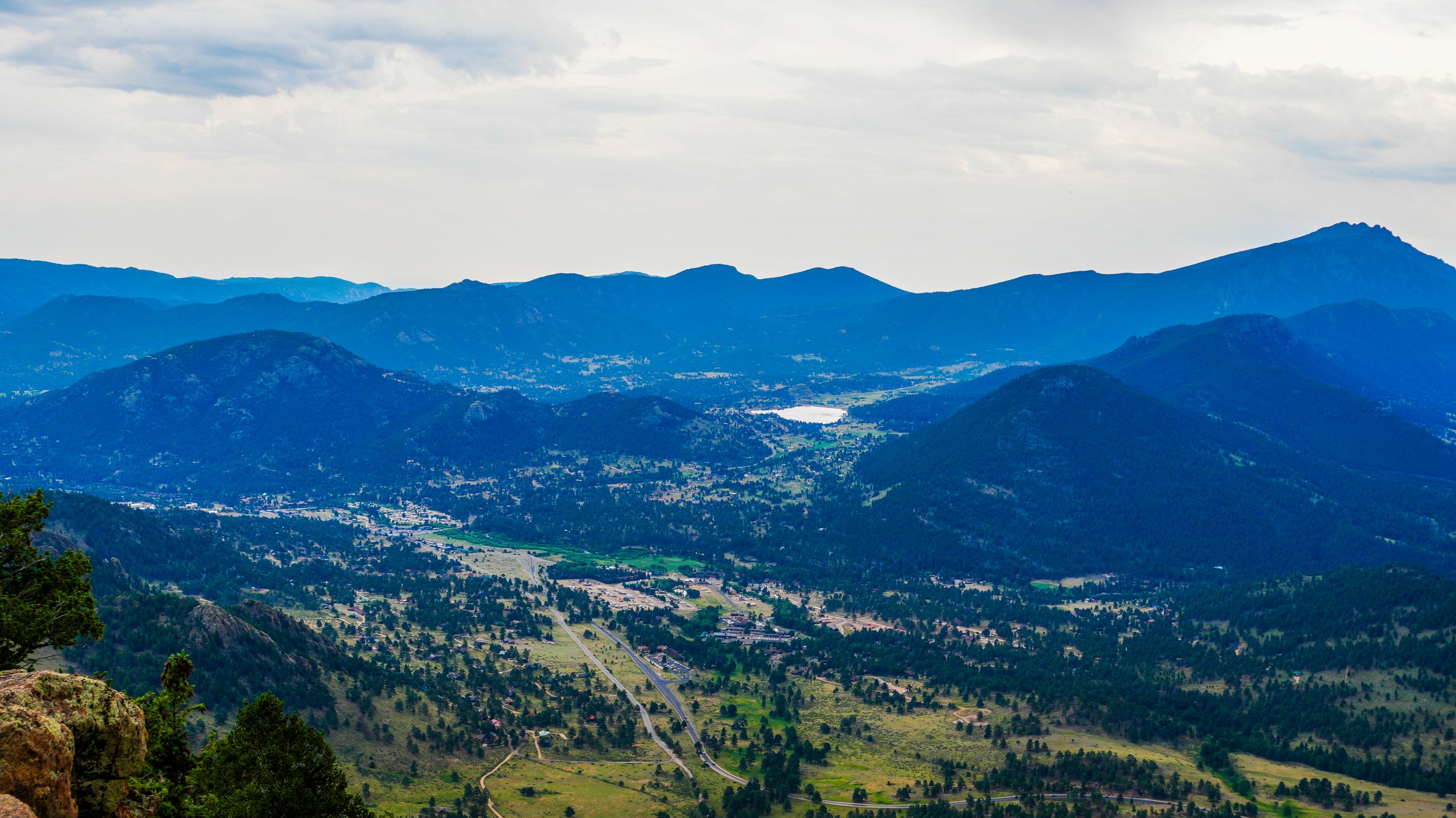 Scenic view of rolling green mountains with a valley and a lake, under a cloudy sky.