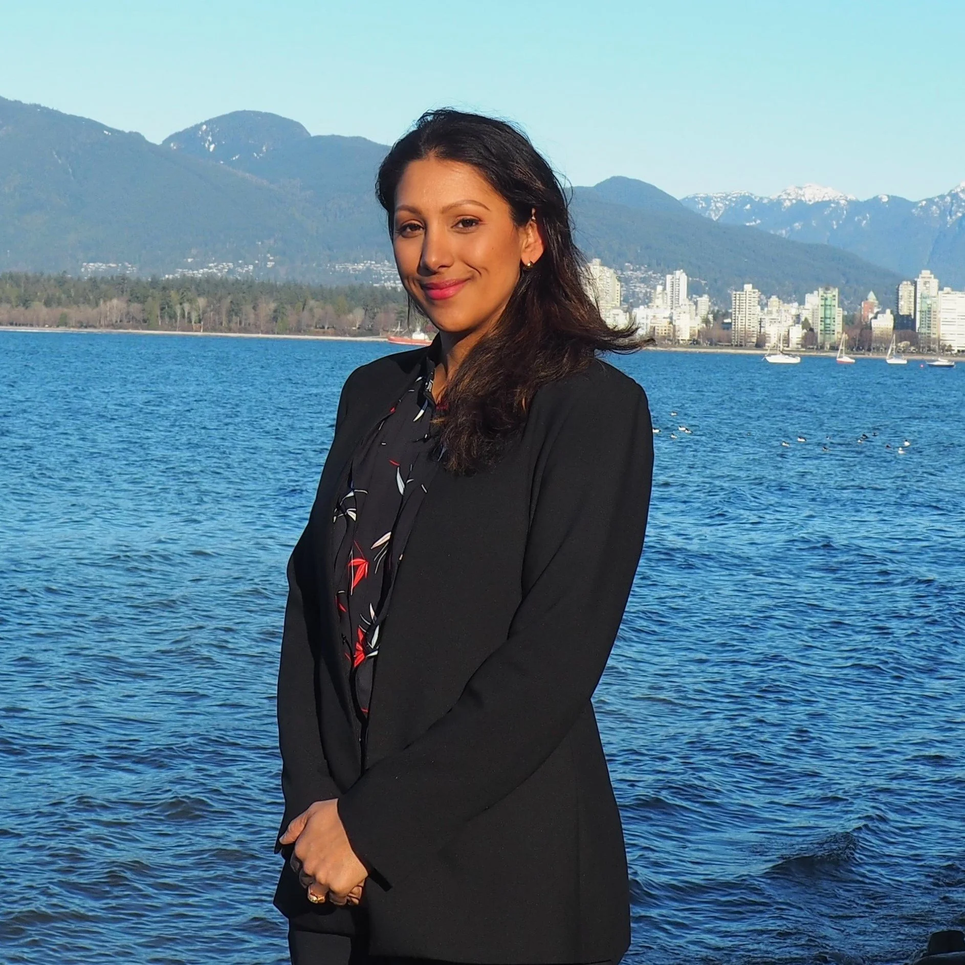 A woman in a black suit standing by a waterfront with mountains and city skyline in the background on a clear day.