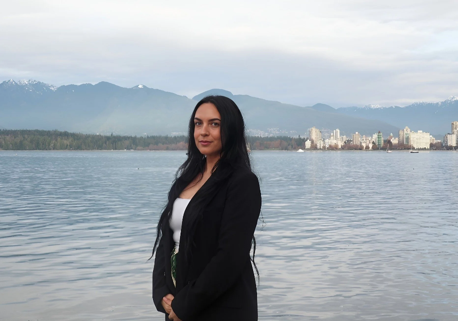 A woman with long black hair standing by a body of water with a city skyline and mountain range in the background.