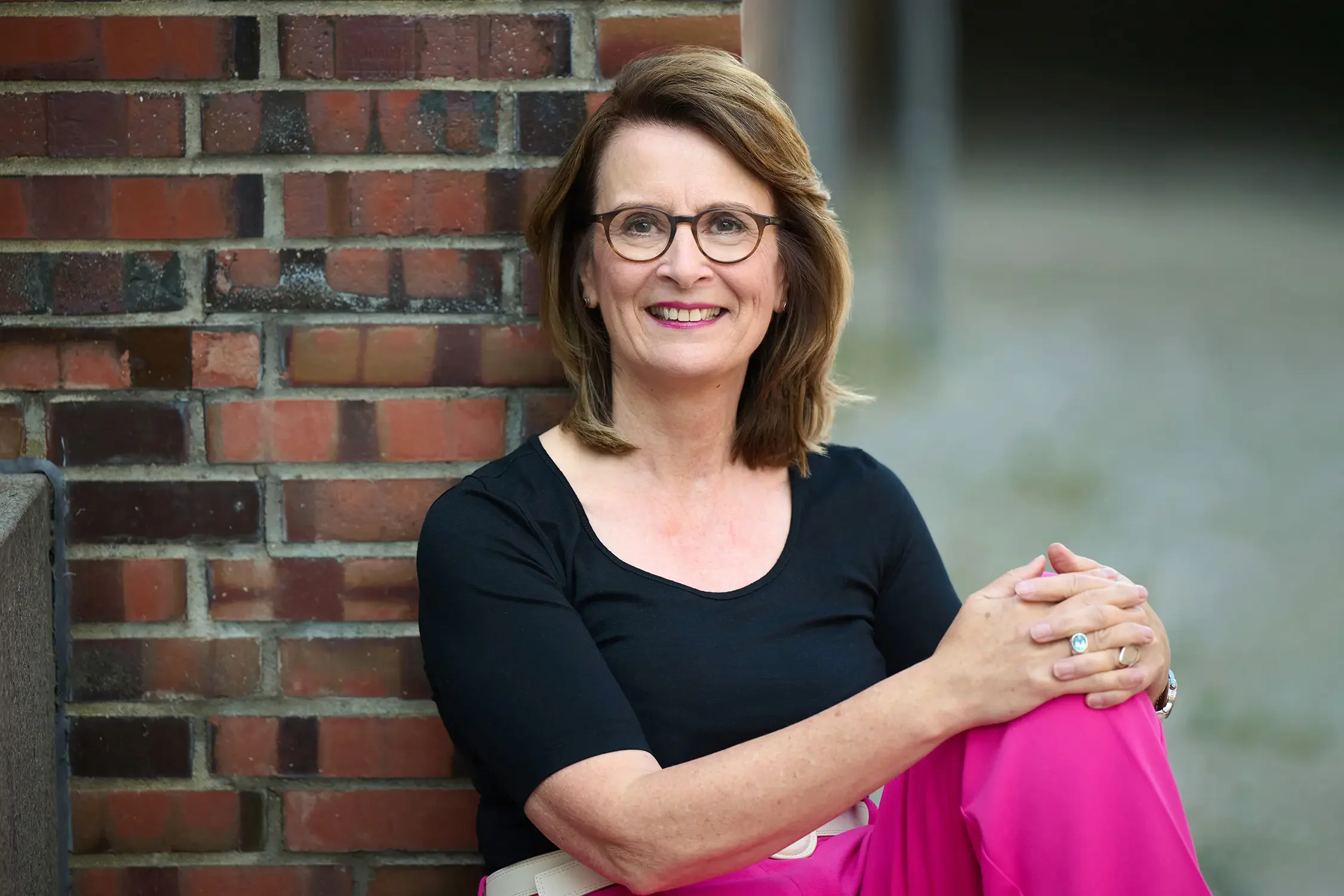 Eine lächelnde Brigitta Wurnig mit Brille sitzt an einer Brickmauer, trägt eine schwarze Oberteil und pinkfarbige Hosen.