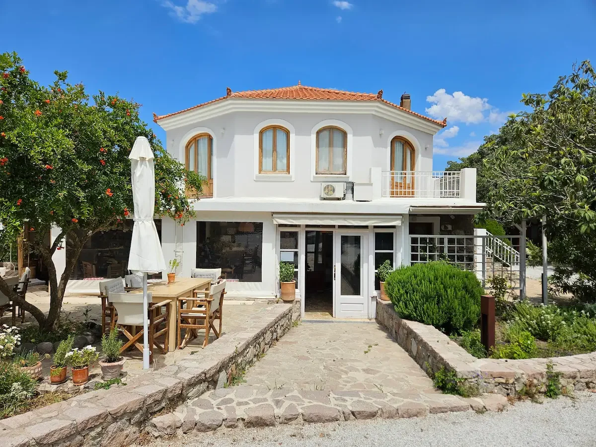 Das weiße Haupthaus von Milelia Inselgarten mit rotem Ziegeldach, umgeben von Gartenpflanzen, mit einem gepflasterten Weg und einer Terrasse mit Tisch und Stühlen im Freien.
