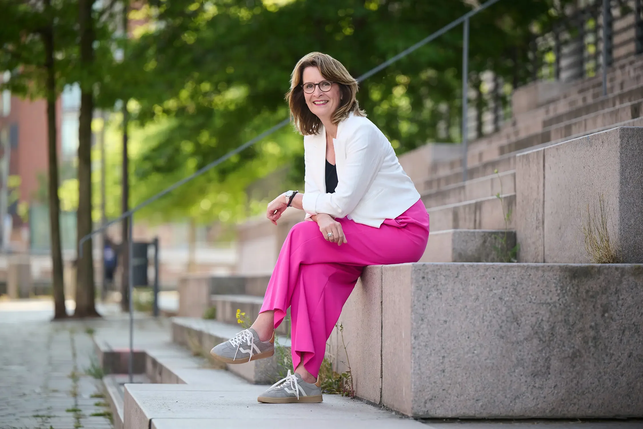 Brigitta Wurnig sitzt auf einer Betonbank im Freien, lächelt und trägt eine weiße Jacke, schwarze Top, pinke Hosen, graue Turnschuhe, Brille und ein Armband.