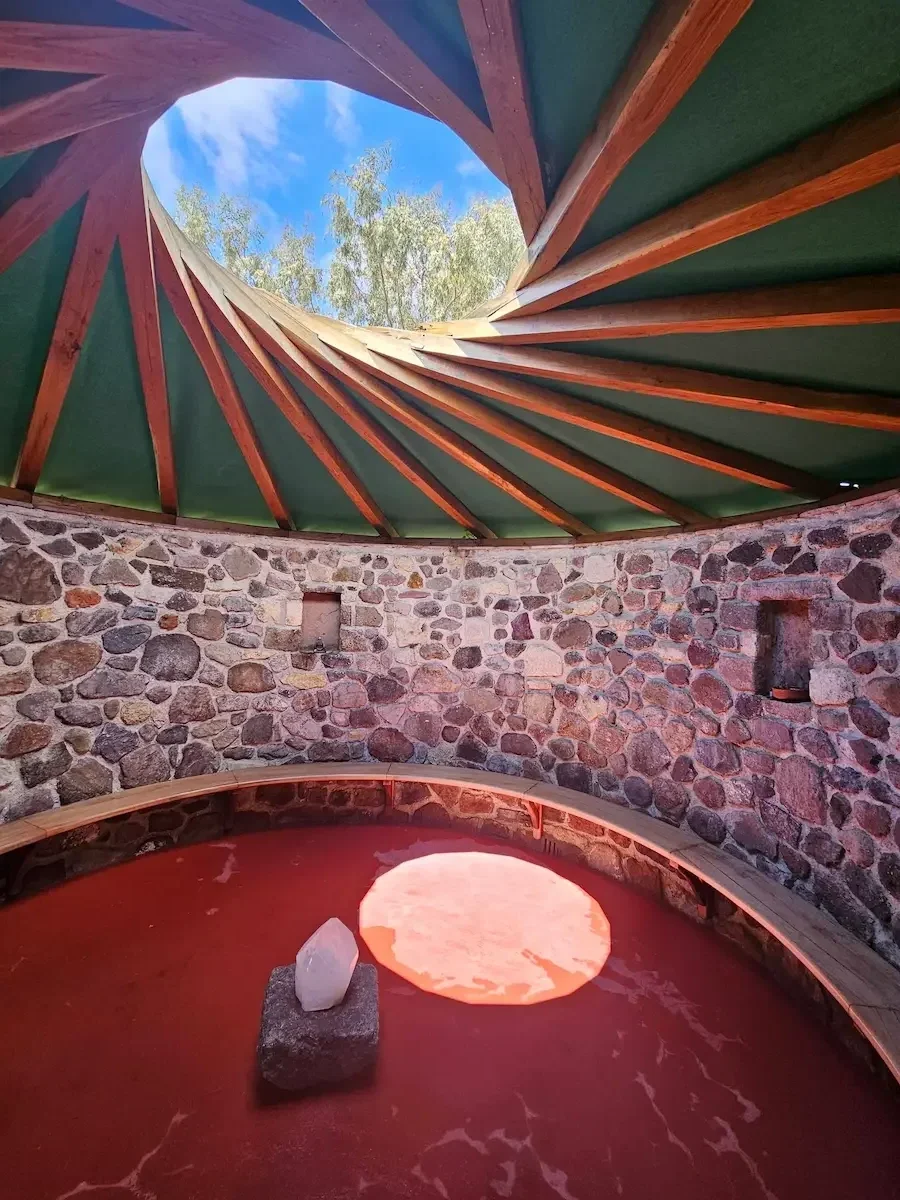 Ein runder Raum mit einer Steinmauer und einem Wasserbecken. Über dem Becken ist ein spitz zulaufender, spiralförmiger Dachüberhang mit grüner Abdeckung und Holzträgern, durch den der blaue Himmel mit Wolken sichtbar ist.
