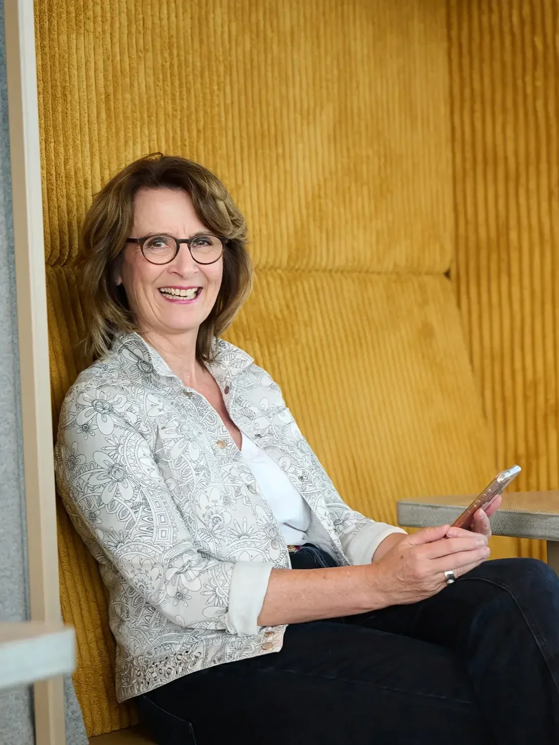 Eine lachende Brigitta Wurnig mit Brille sitzt auf einer grünen Bank neben einer Wand mit einem grünen und orangenen Muster und hält ein Smartphone in der Hand.