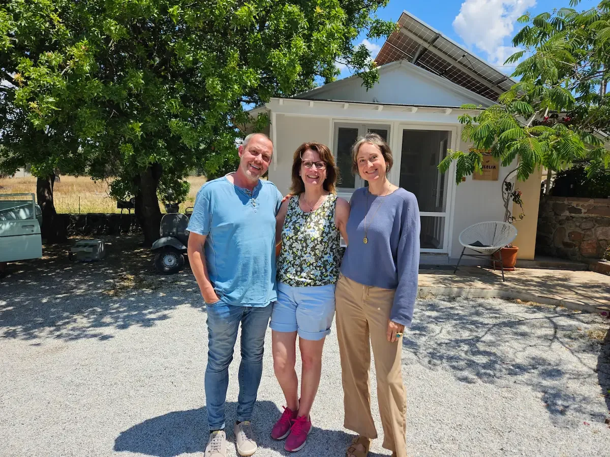 Brigitta Wurnig steht mit zwei Menschen vor einem weißen Haus im Milelia Inselgarten mit Garten im Hintergrund. Es ist sonnig, einige Bäume spenden Schatten, und die drei Personen lachen und posieren für das Foto.