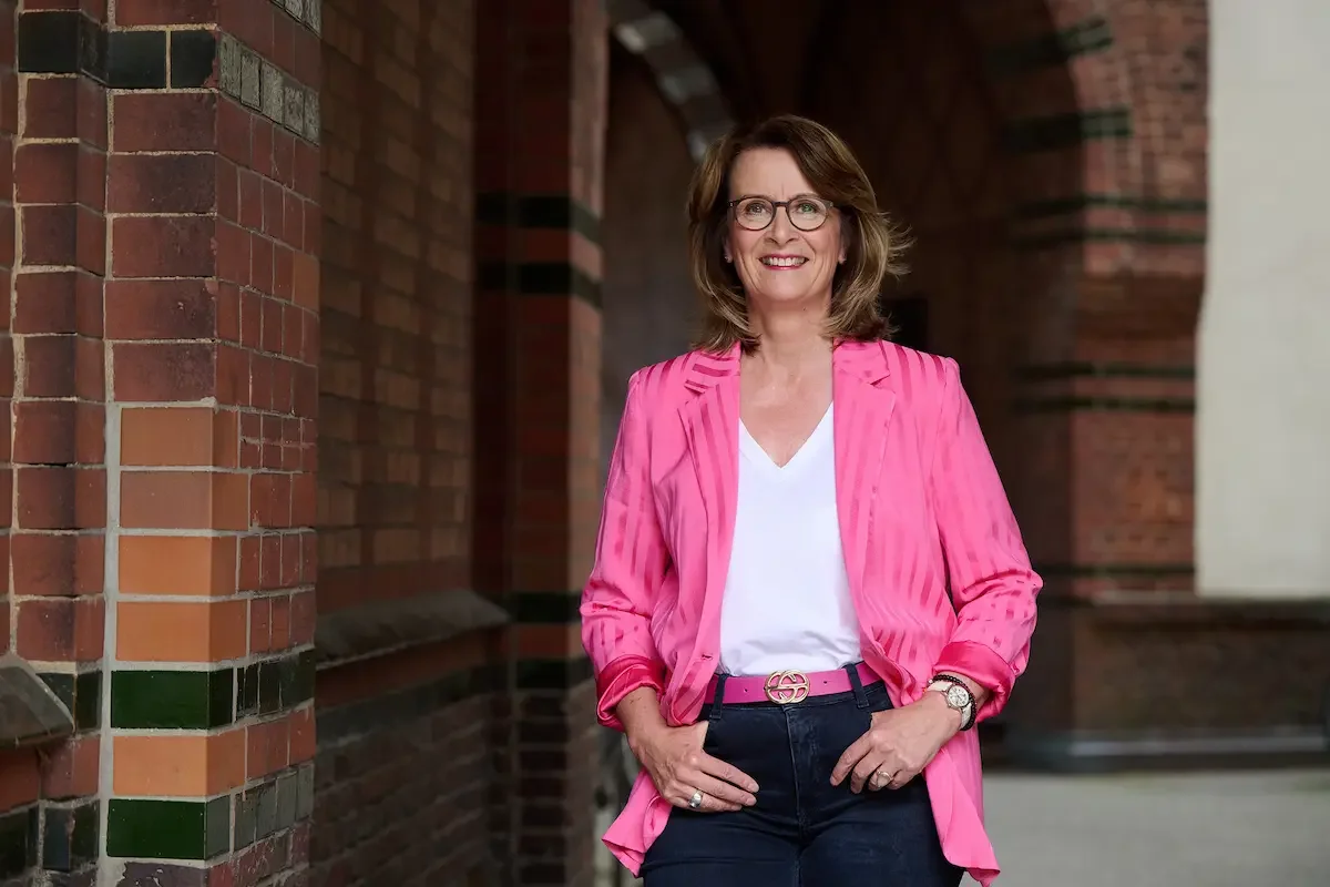 Brigitta Wurnig mit Brille, braunen Haaren und einem Lächeln, die in einem pinken Blazer, weißem T-Shirt und dunklen Hosen vor einer Backsteinmauer steht.