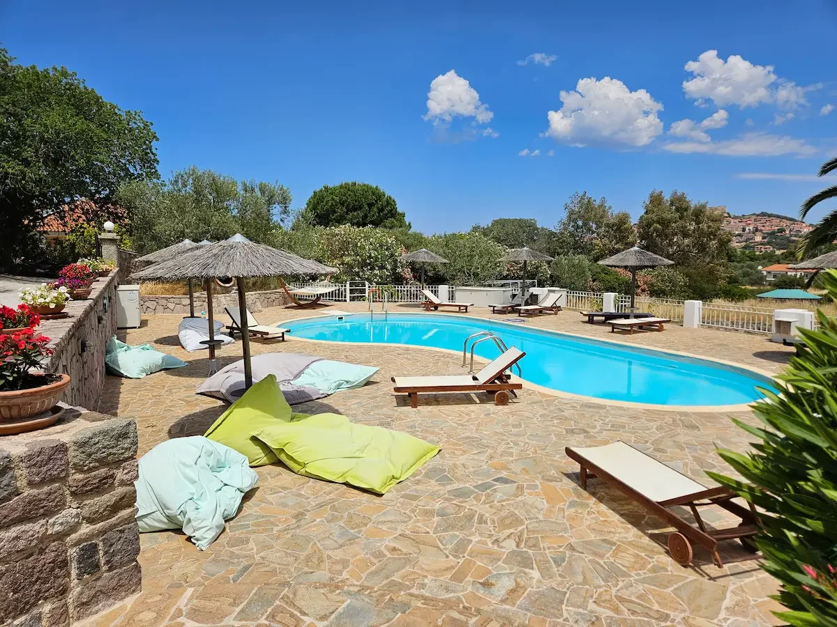 Der Poolbereich von Milelia Inselgarten im Freien mit Liegestühlen, Sonnenschirmen, Blumenkübeln, gepolsterten Sitzsäcken und einer Solardusche bei sonnigem Himmel.