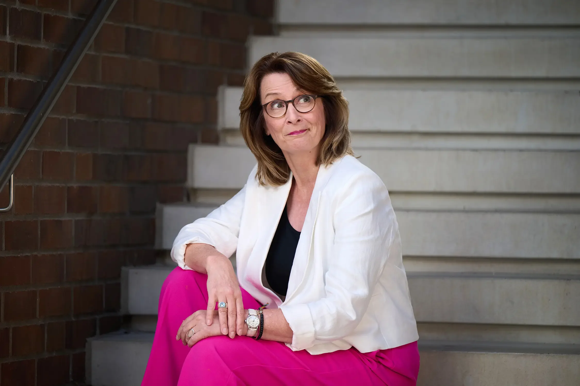 Brigitta Wurnig mit Brille in einem weißen Jacke und pinken Hosen sitzt auf Stufen und schaut nach oben.