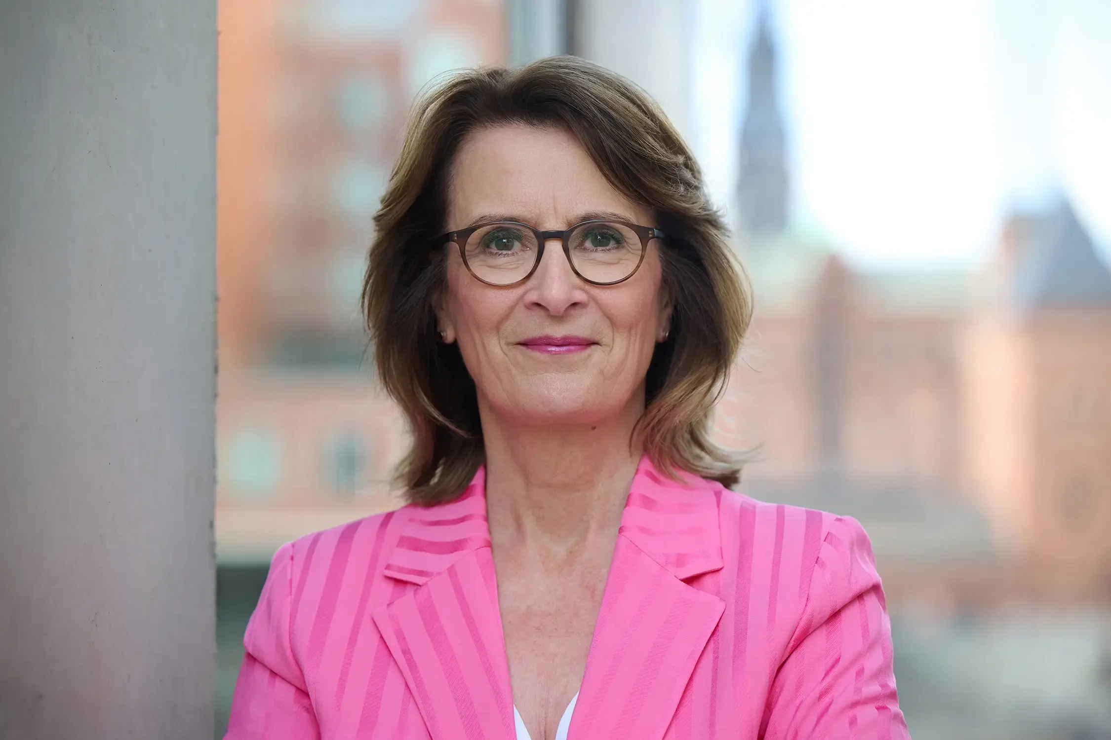 Brigitta Wurnig mit Brille in pinkem Blazer vor einem Fenster, unscharfer Städtehintergrund.