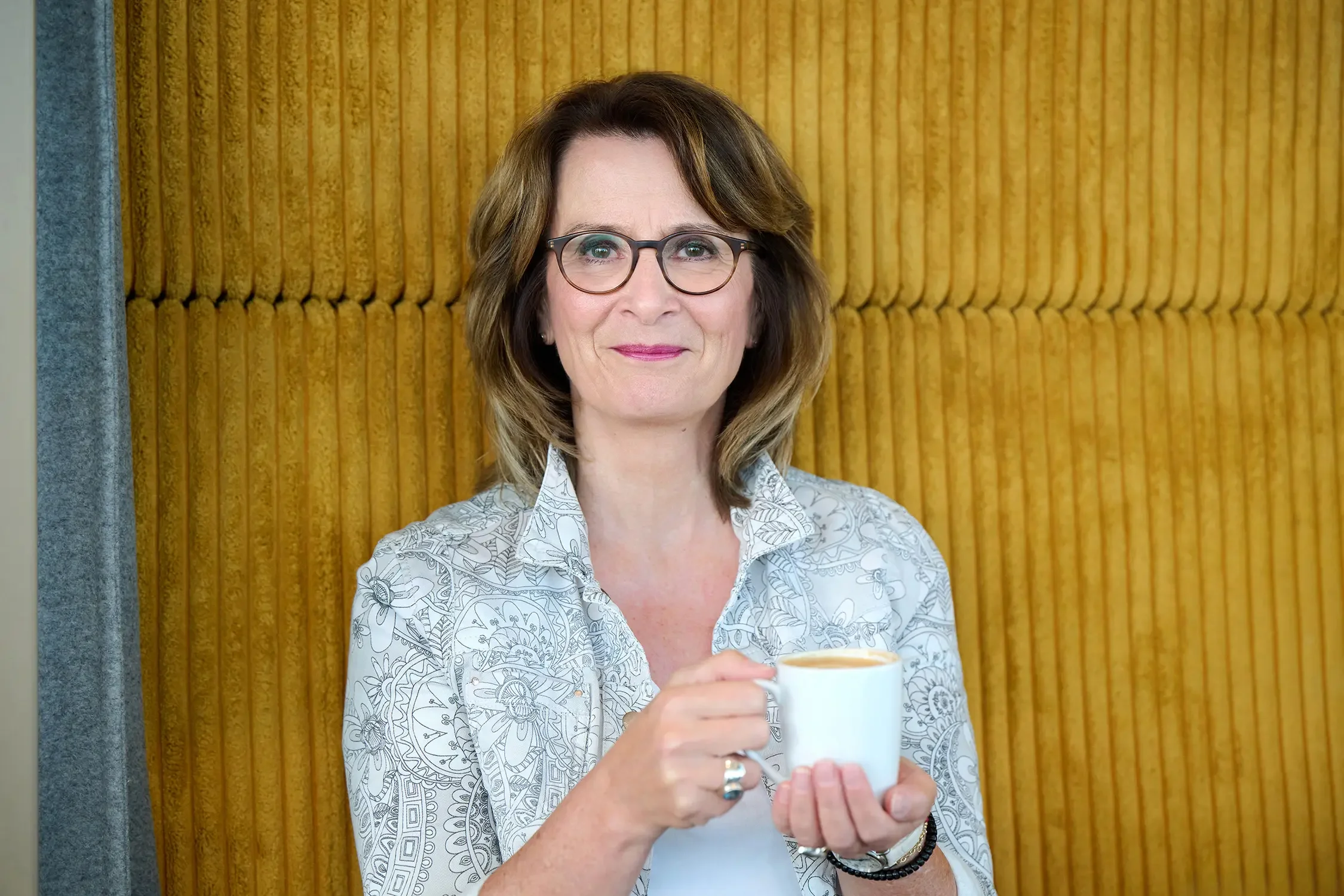 Eine lächelnde Brigitta Wurnig mit Brille hält eine Tasse Kaffee in einer Hand, vor einem gelben gepolsterten Hintergrund.
