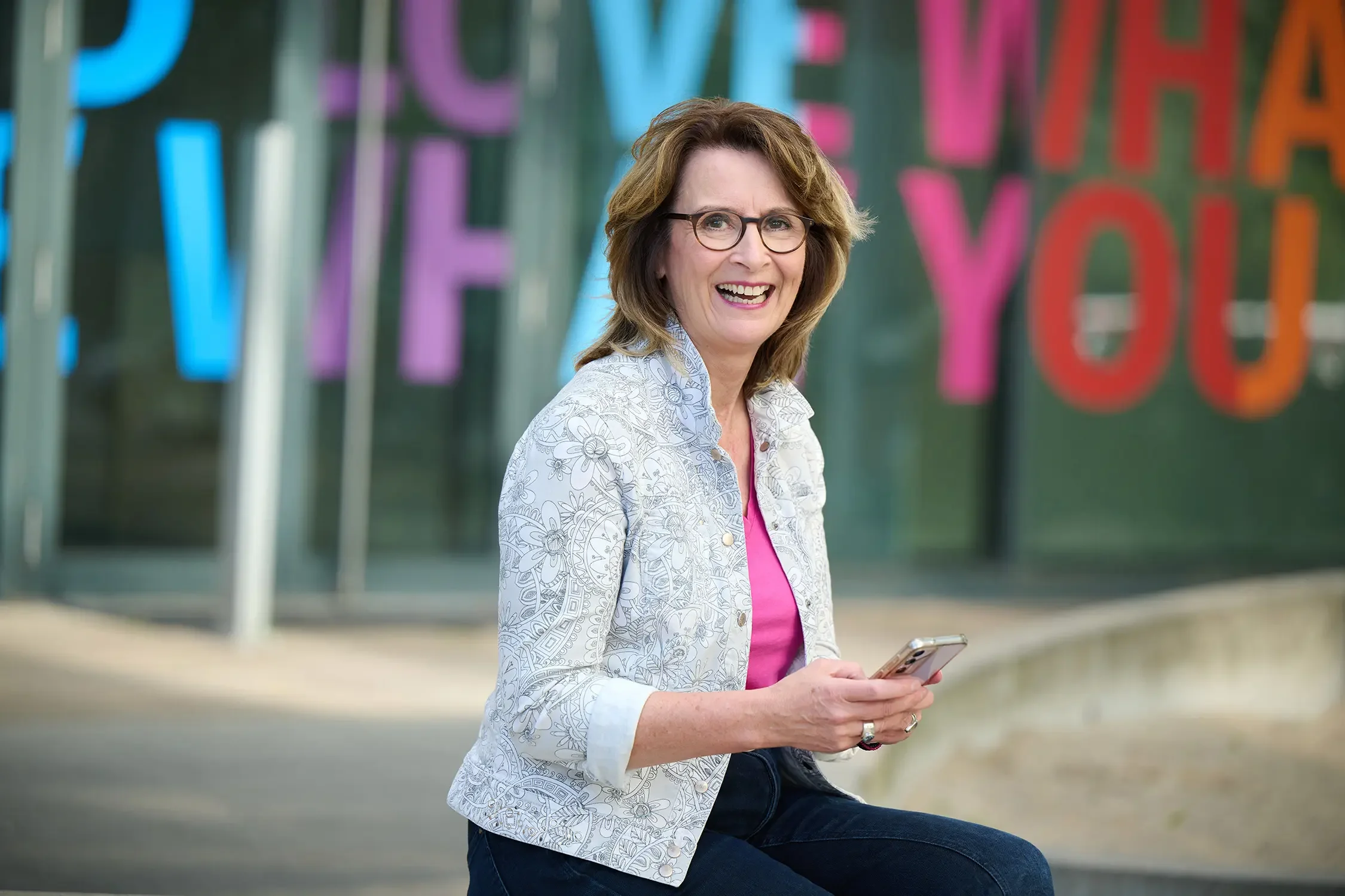 Eine lachende Brigitta Wurnig mit Brille sitzt draußen vor einem Gebäude mit bunten Buchstaben im Hintergrund, hält ein Smartphone in der Hand.